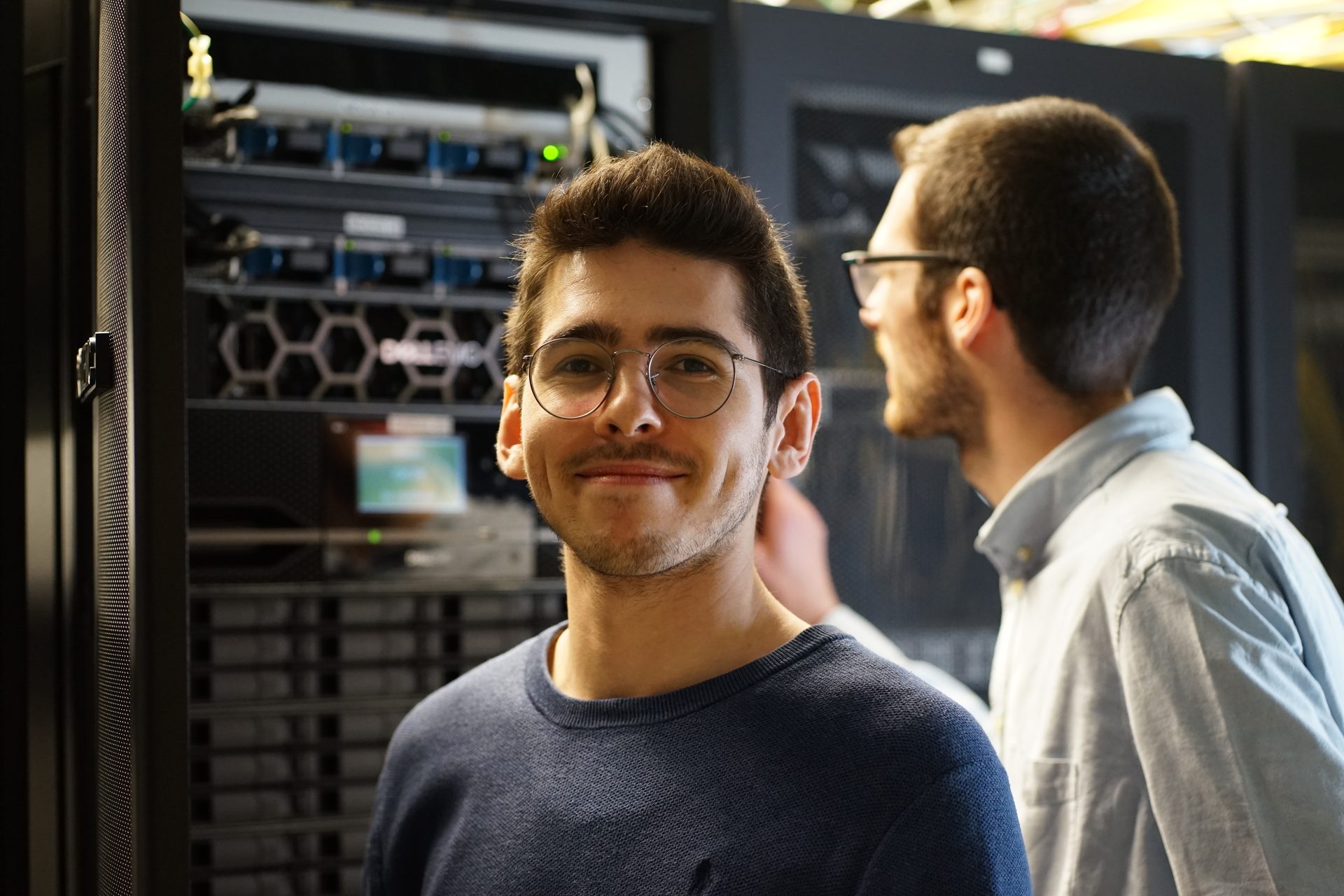 Smiling man in glasses in a server room, with another person partially visible behind him.