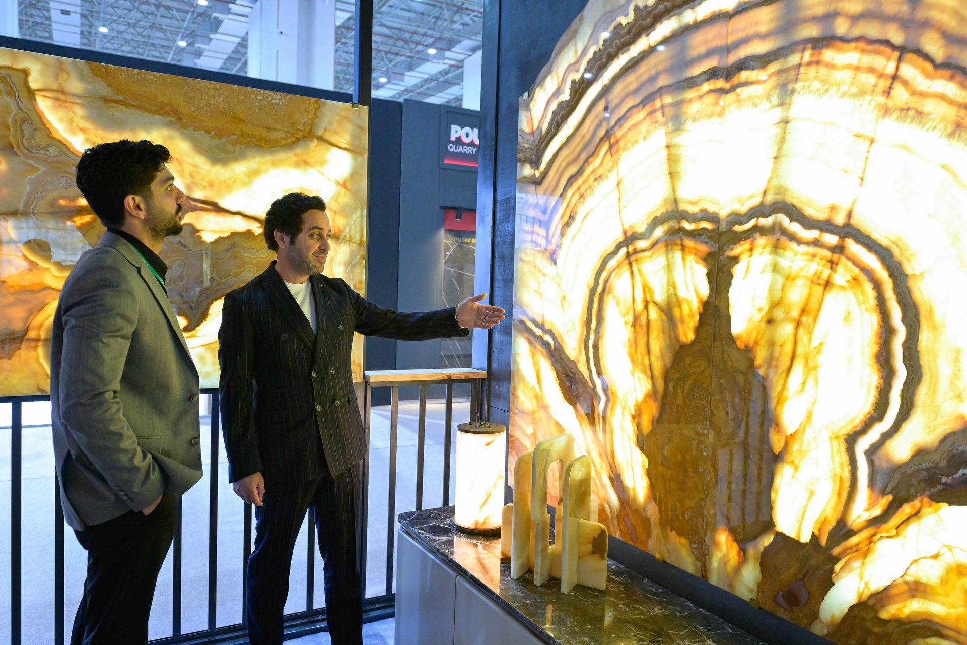 Two men discuss a large, brightly backlit onyx wall panel with unique patterns at an exhibition.