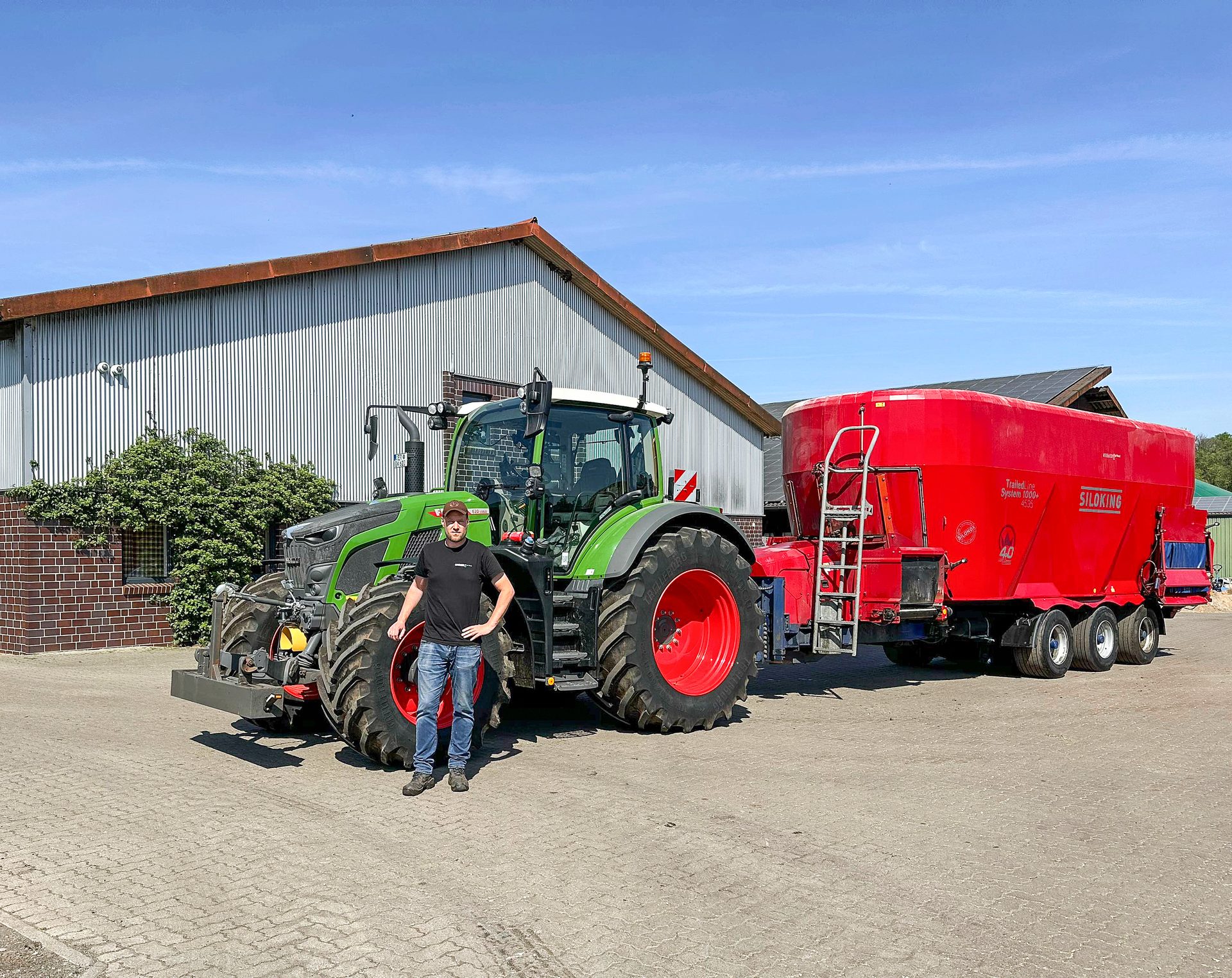Man poses with a green Fendt tractor and red Siloking feeder wagon in front of a farm building.