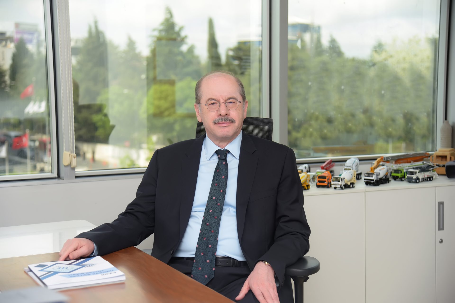 Man in a suit sits at an office desk with miniature construction vehicles and a window view.