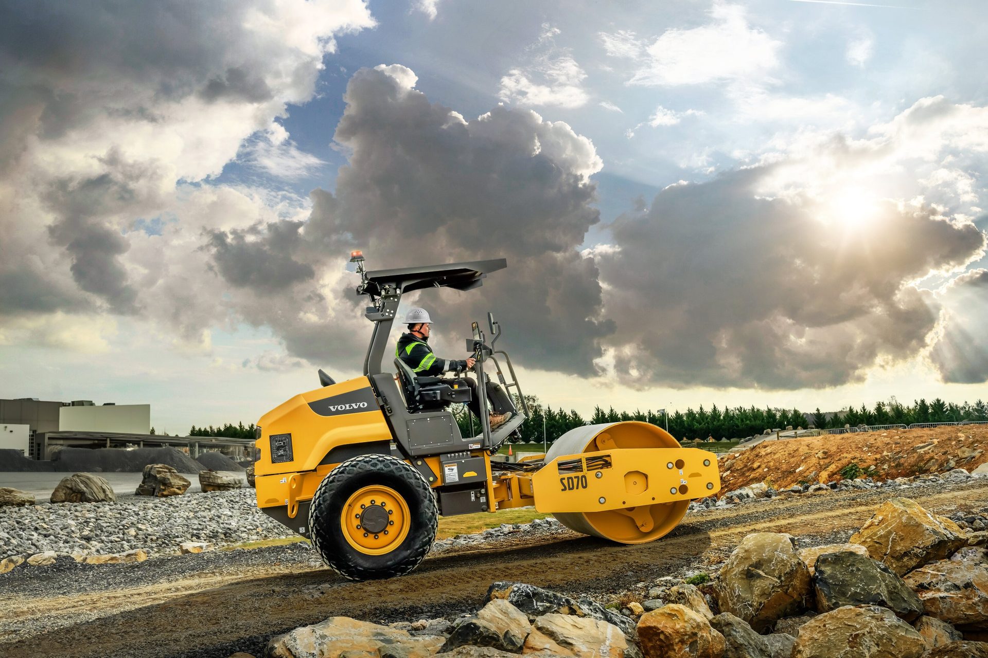 A yellow Volvo SD70 road roller with an operator compacting ground on a rocky construction site.
