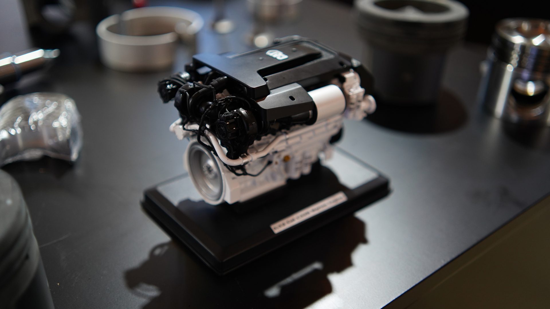 Miniature white and black engine model on a black stand, surrounded by blurred metallic objects.