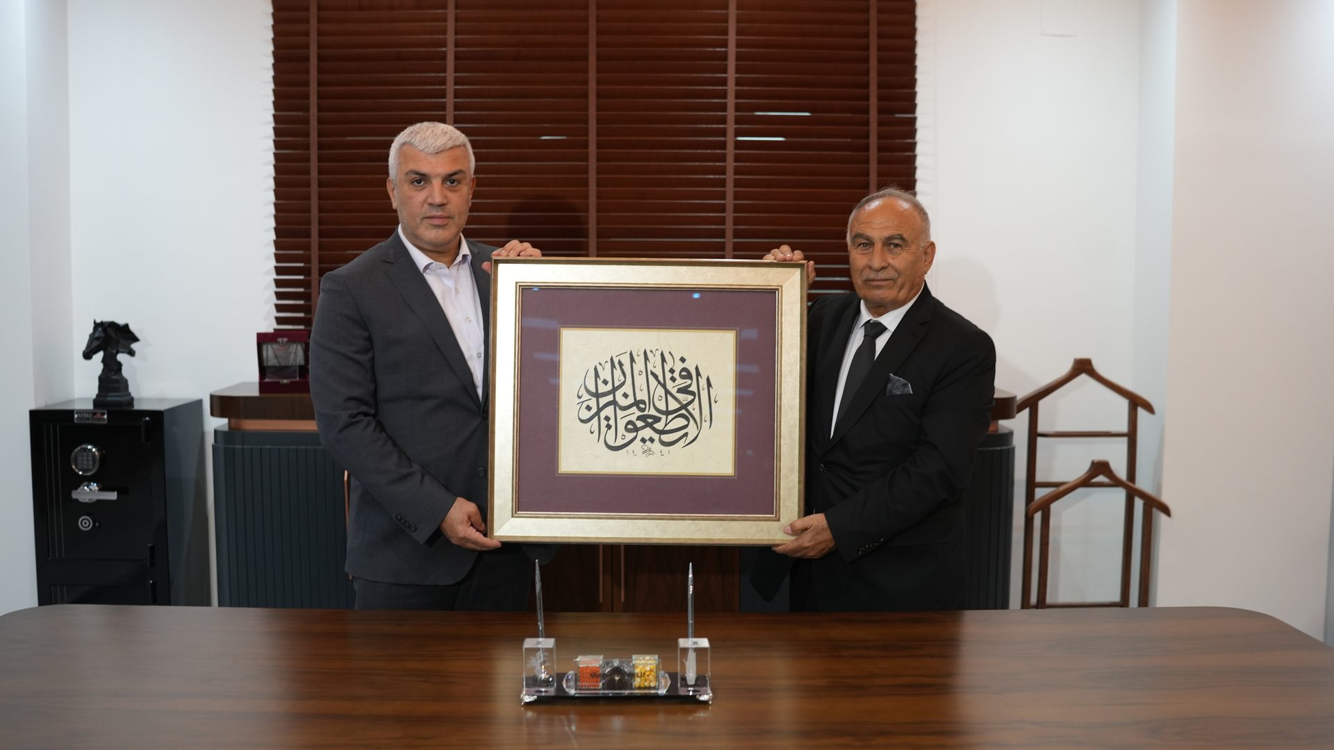 Two men in suits holding a framed artwork with Arabic calligraphy.