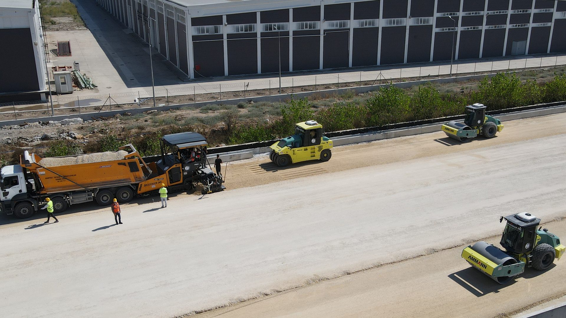 Road paving in progress: dump truck, paver, and rollers on a new roadbed with workers.