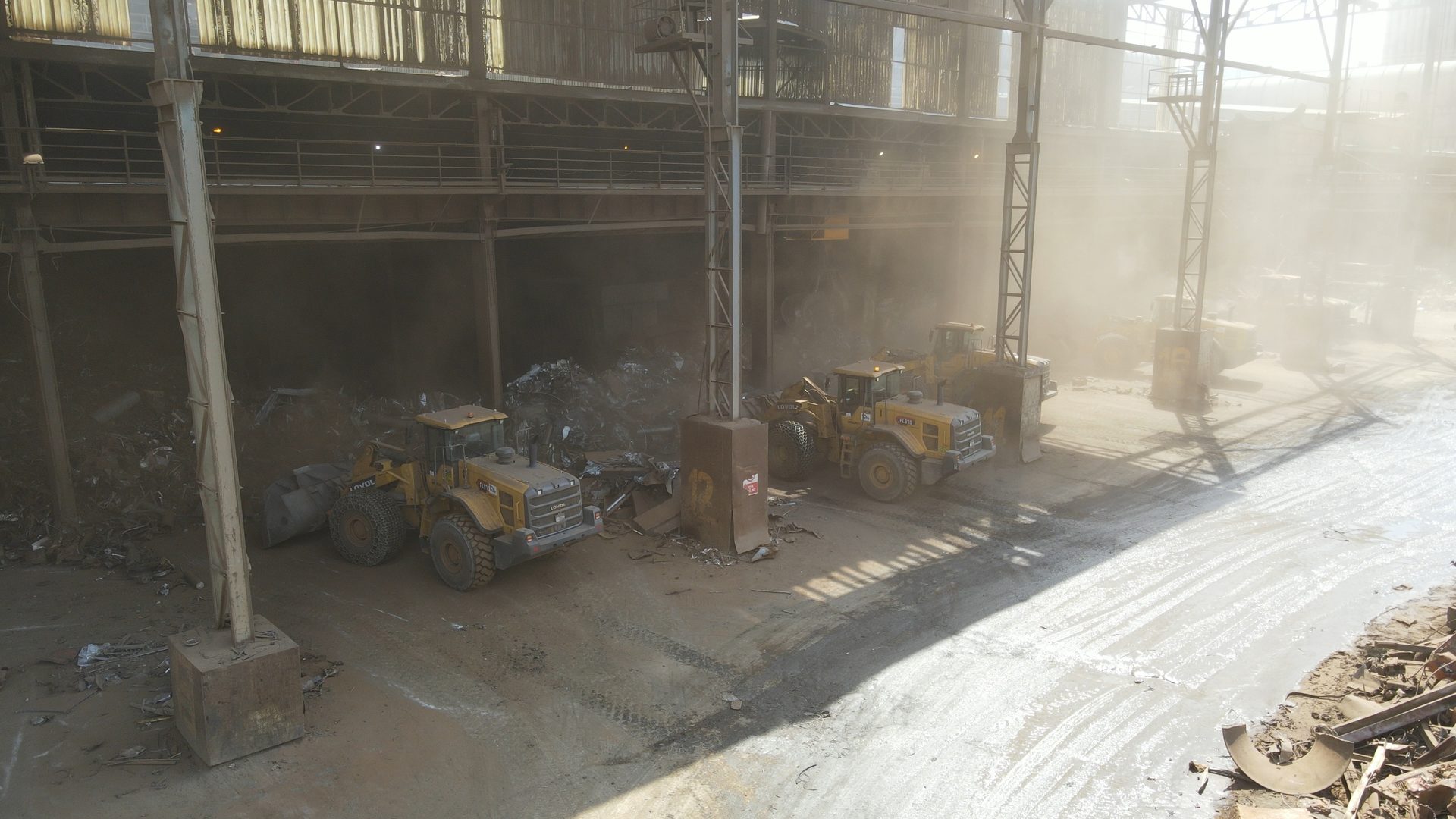 Yellow loaders moving scrap metal in a dusty industrial facility.