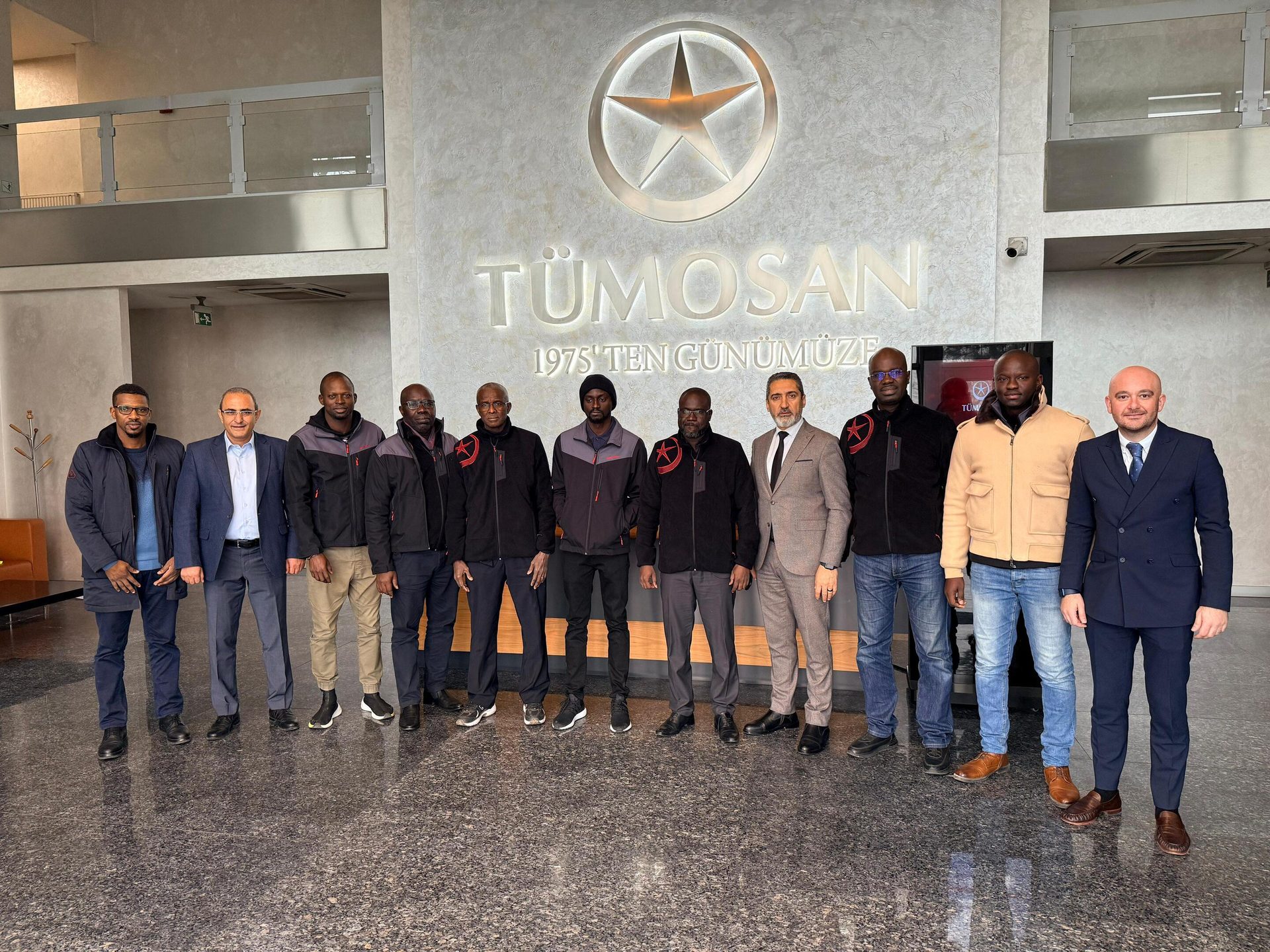 Eleven men, some in suits and some in casual attire, pose in front of a "TÜMOSAN" sign.