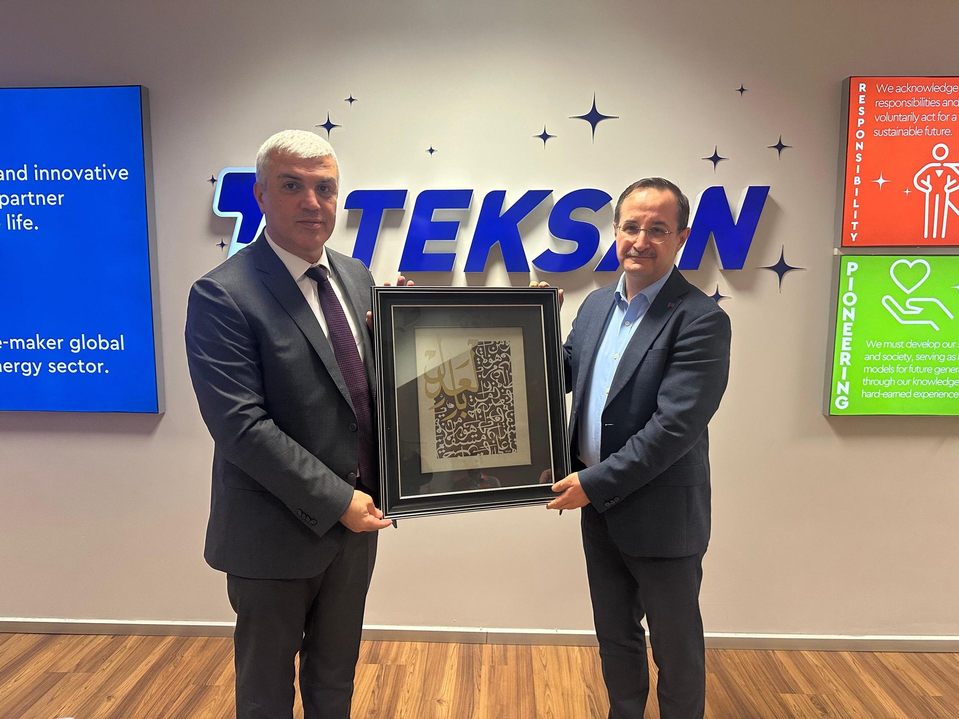Two men in suits hold a framed calligraphic artwork in a TEKSON office.