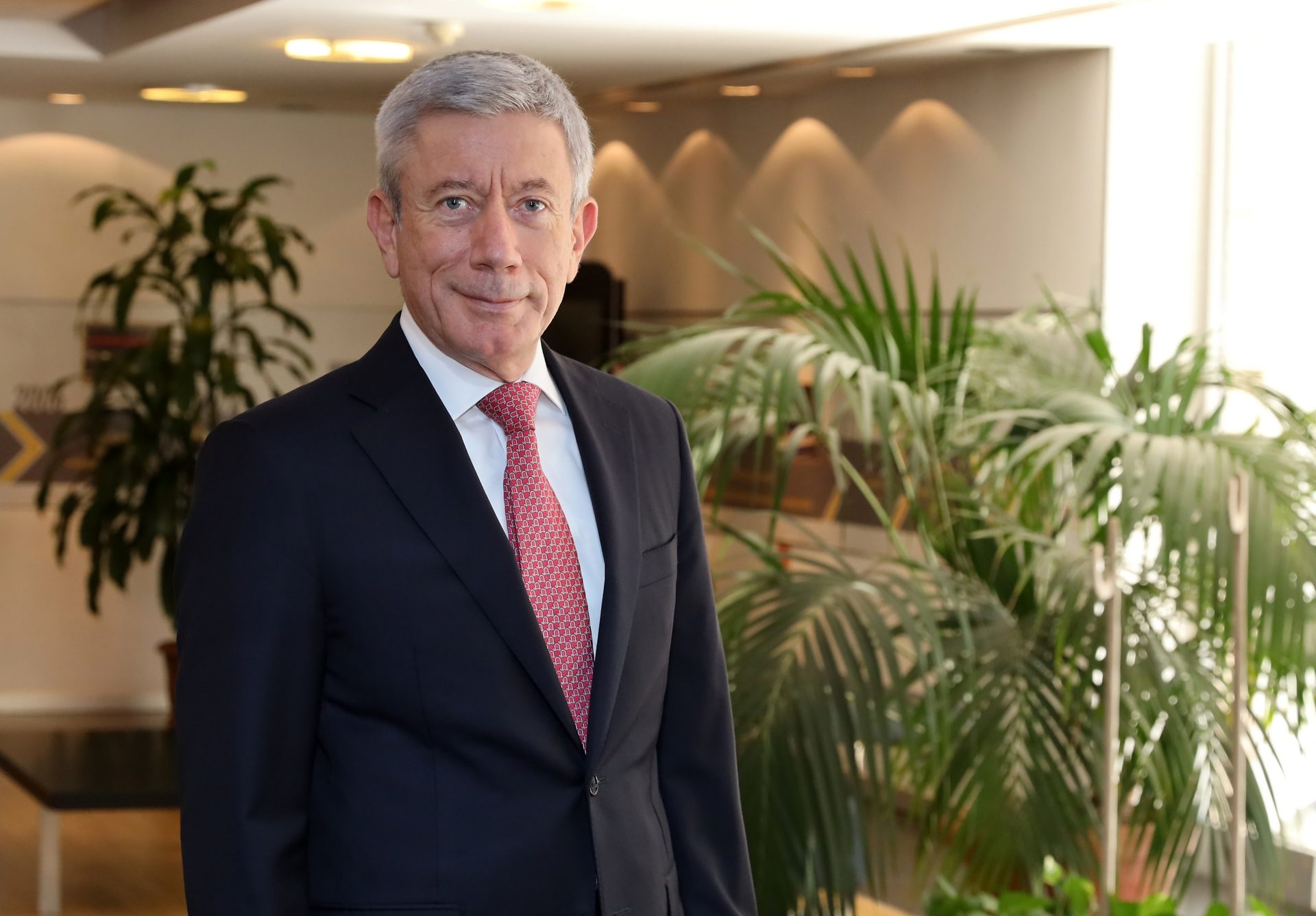 Smiling man with grey hair in a dark suit and red tie, in an office with plants.