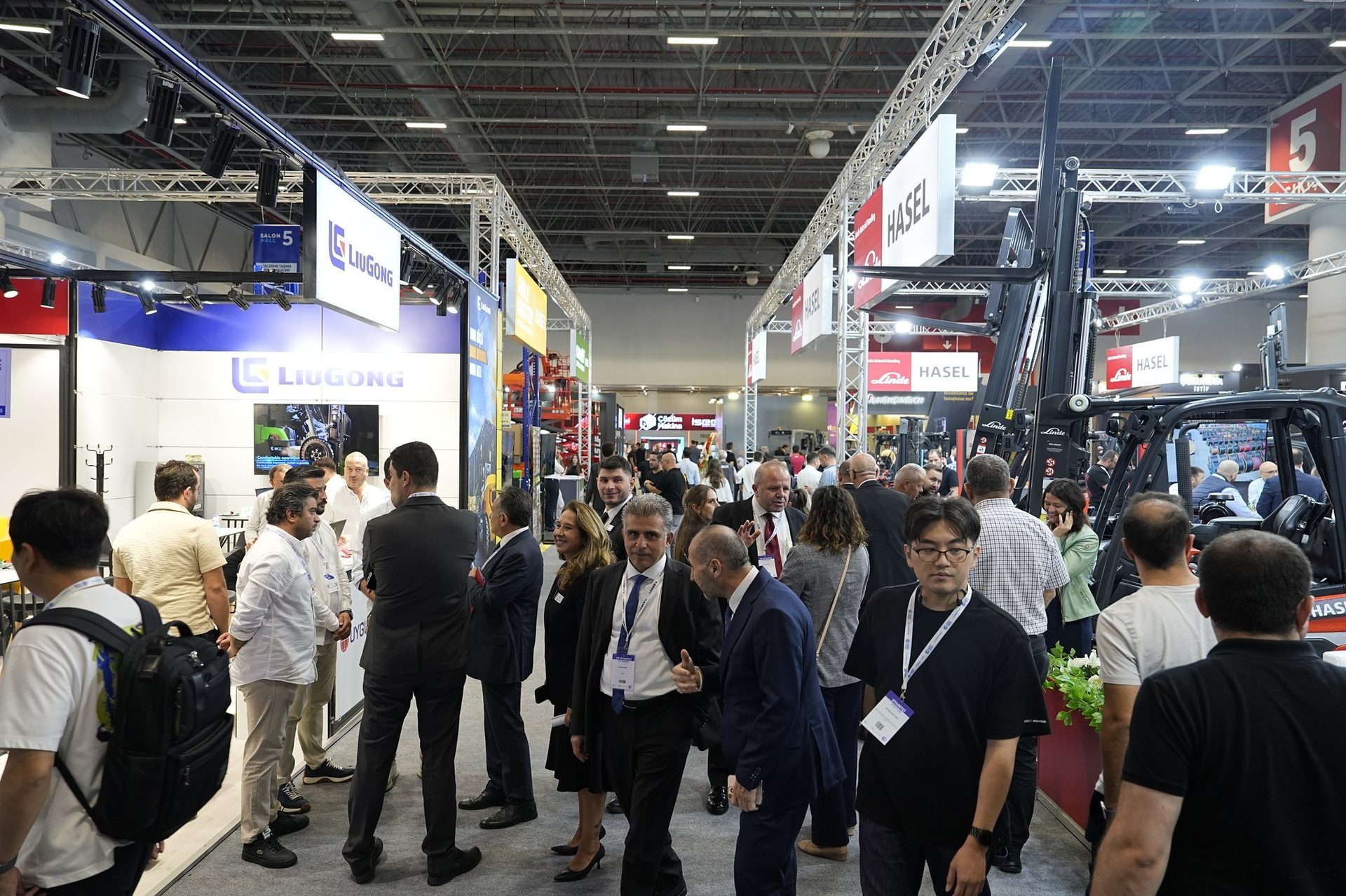 Bustling trade show with attendees, LIUGONG and HASEL booths, and forklifts visible.