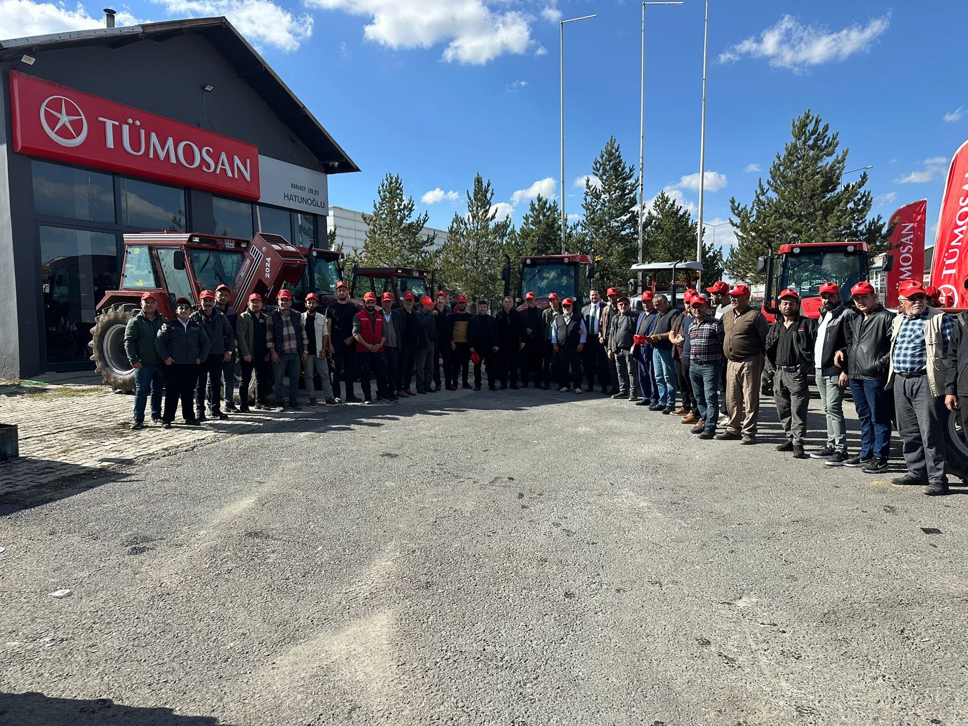 A group of men in red hats poses in front of a TÜMOSAN building with red tractors.