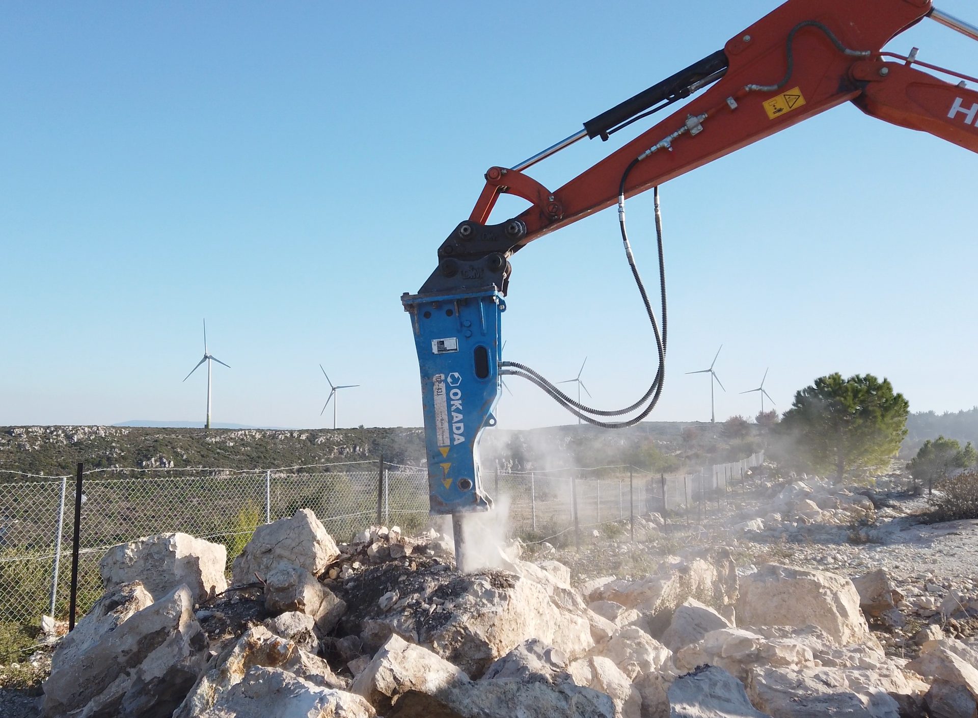 An excavator with a blue hydraulic breaker demolishes rocks, with wind turbines visible in the background.