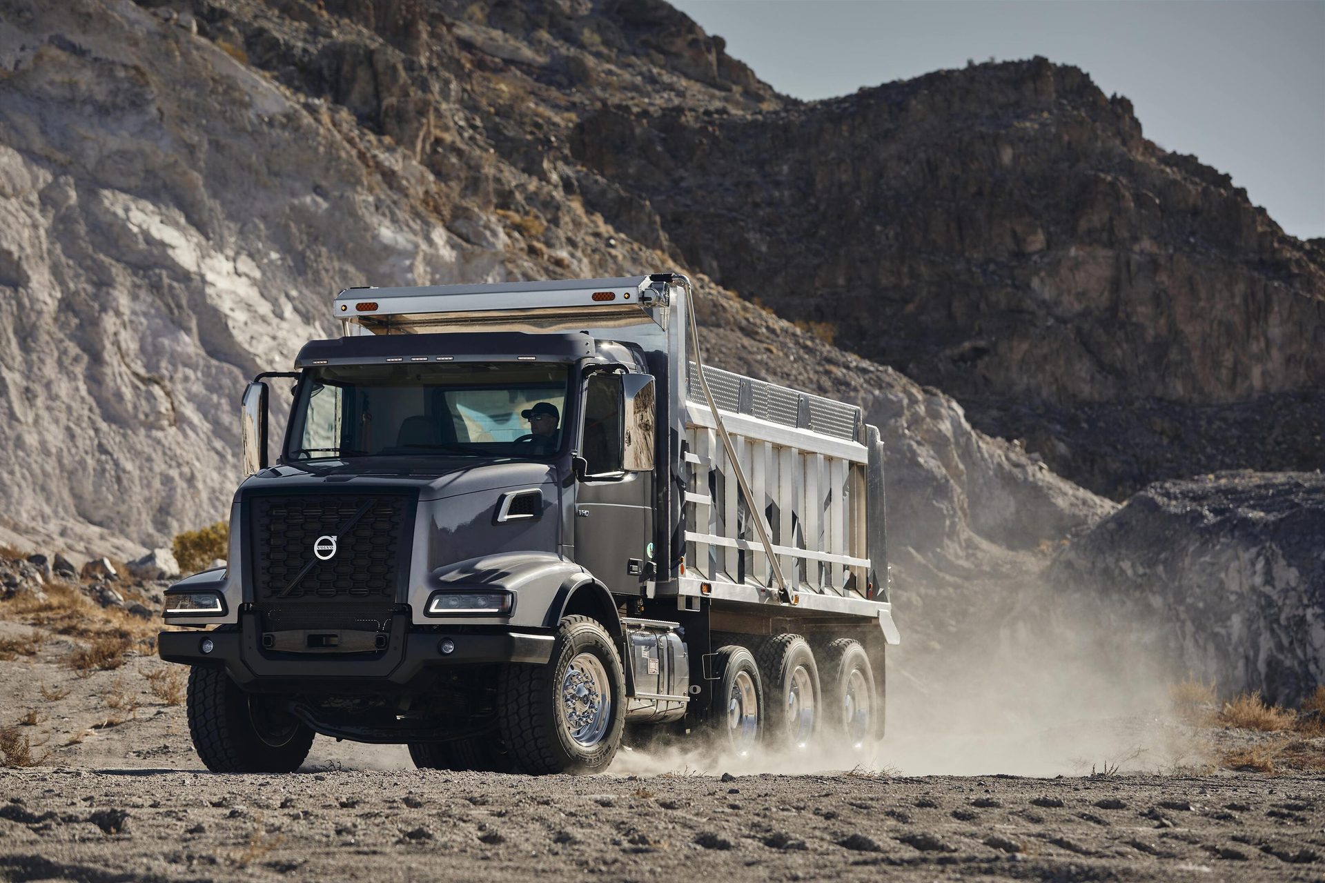 A gray Volvo dump truck drives on a dusty unpaved road in a rocky, mountainous desert landscape, stirring up dust.