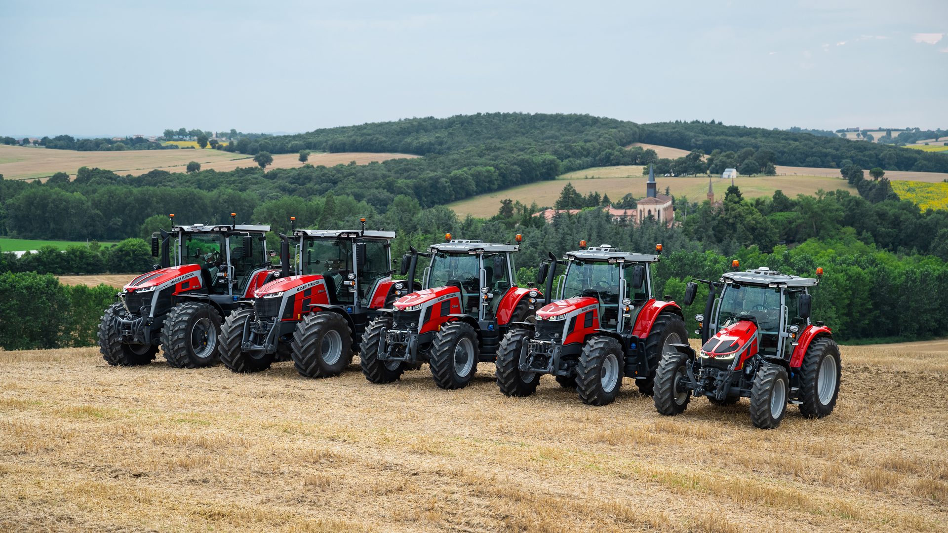 Six red Massey Ferguson tractors lined up in a field, with a village and green hills in the background.