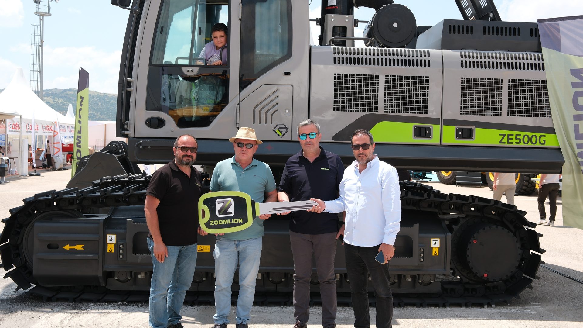 Four men and a child with a large Zoomlion excavator, one man holding a giant key.