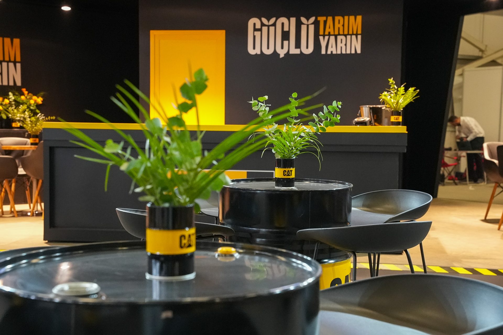 Industrial lounge with oil drum tables, CAT-branded plants, black chairs, and yellow-accented walls.