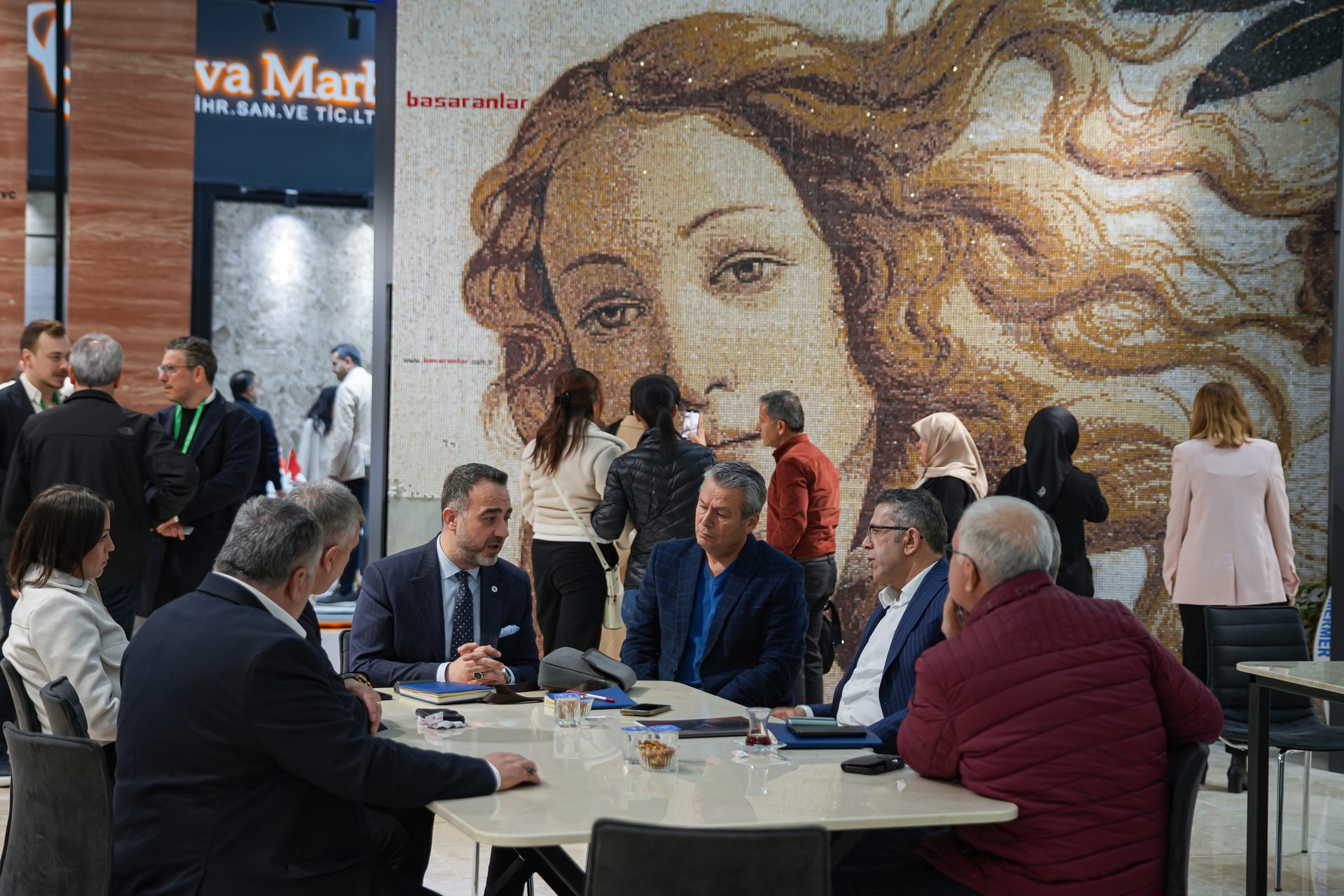 People conversing around a table at an exhibition, with a large mosaic of a woman's face in the background.