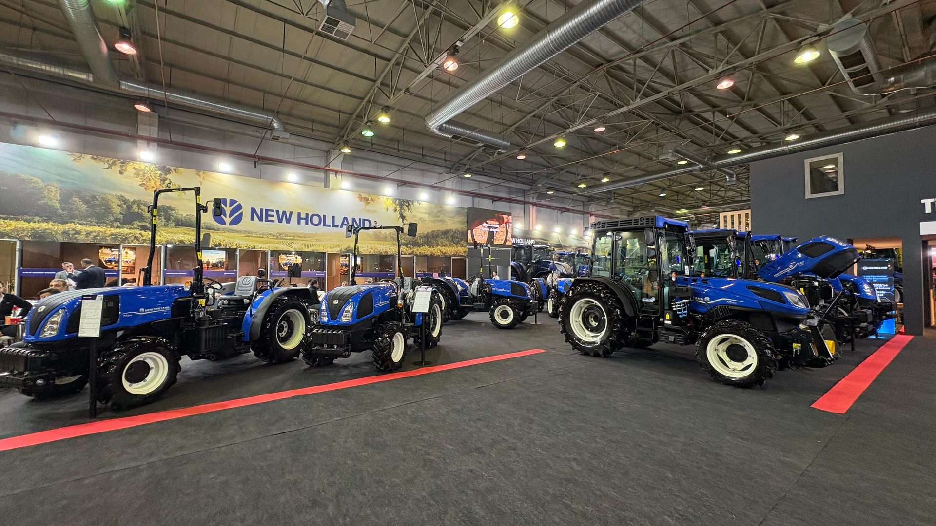 Blue New Holland tractors on display at an indoor trade show, with a large brand banner.
