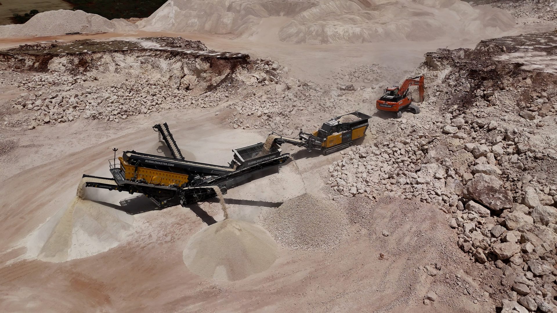 Aerial view of a quarry. An excavator feeds rocks into a crusher, producing aggregate material.
