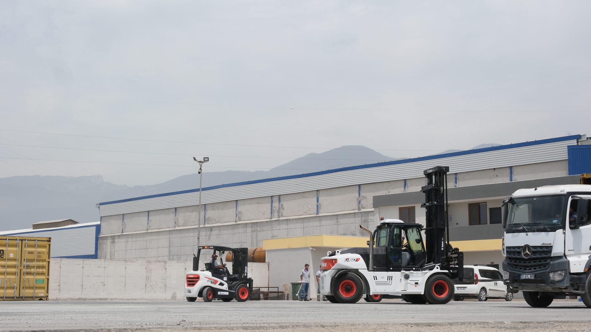Heavy forklifts and truck in a logistics yard with workers, buildings, and mountains.