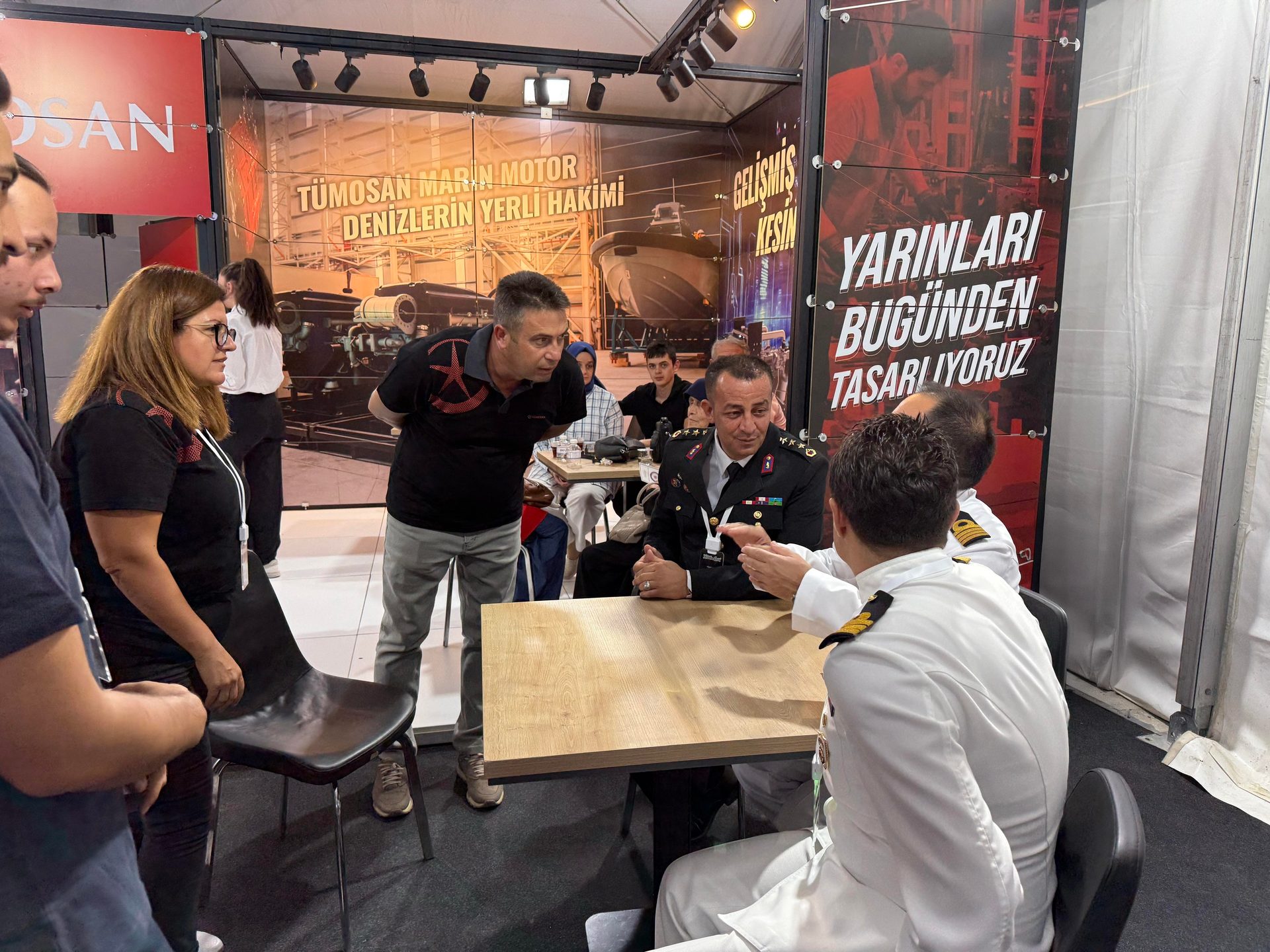 Group of people, including two military personnel, at a booth with marine engine banners.
