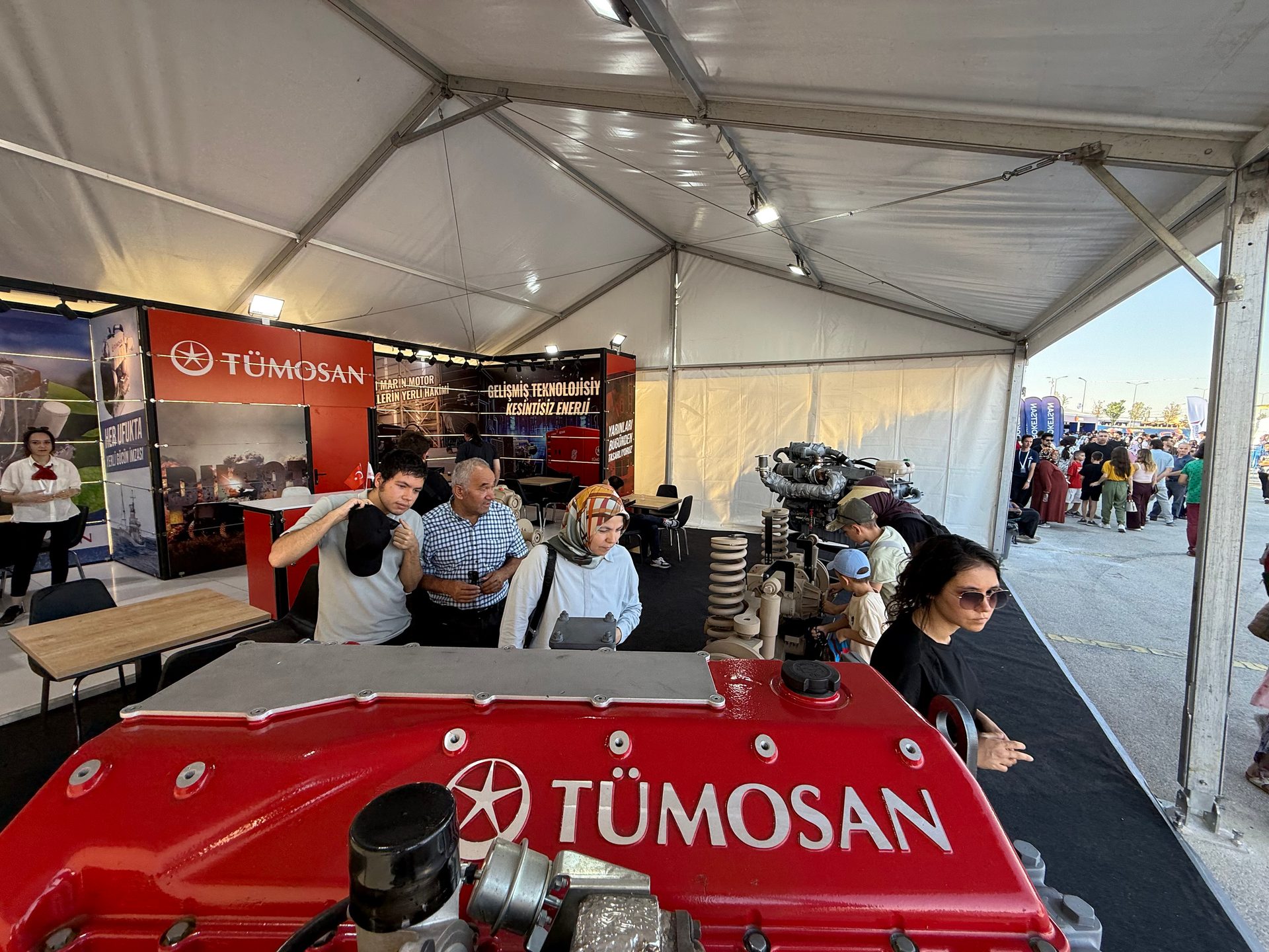 Indoor exhibition booth for Tümosan, featuring a red engine and visitors under a tent.