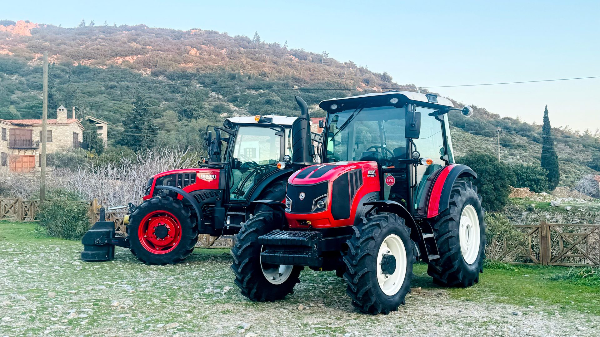 Two red tractors in a rural landscape with hills and a stone house.