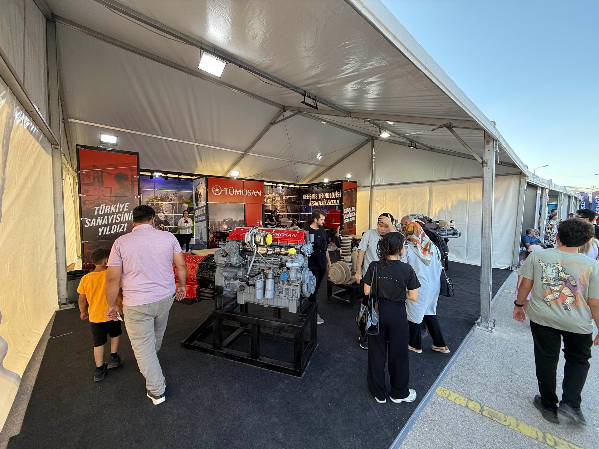 People examining a large industrial engine at a TÜMOSAN exhibit inside a white tent.
