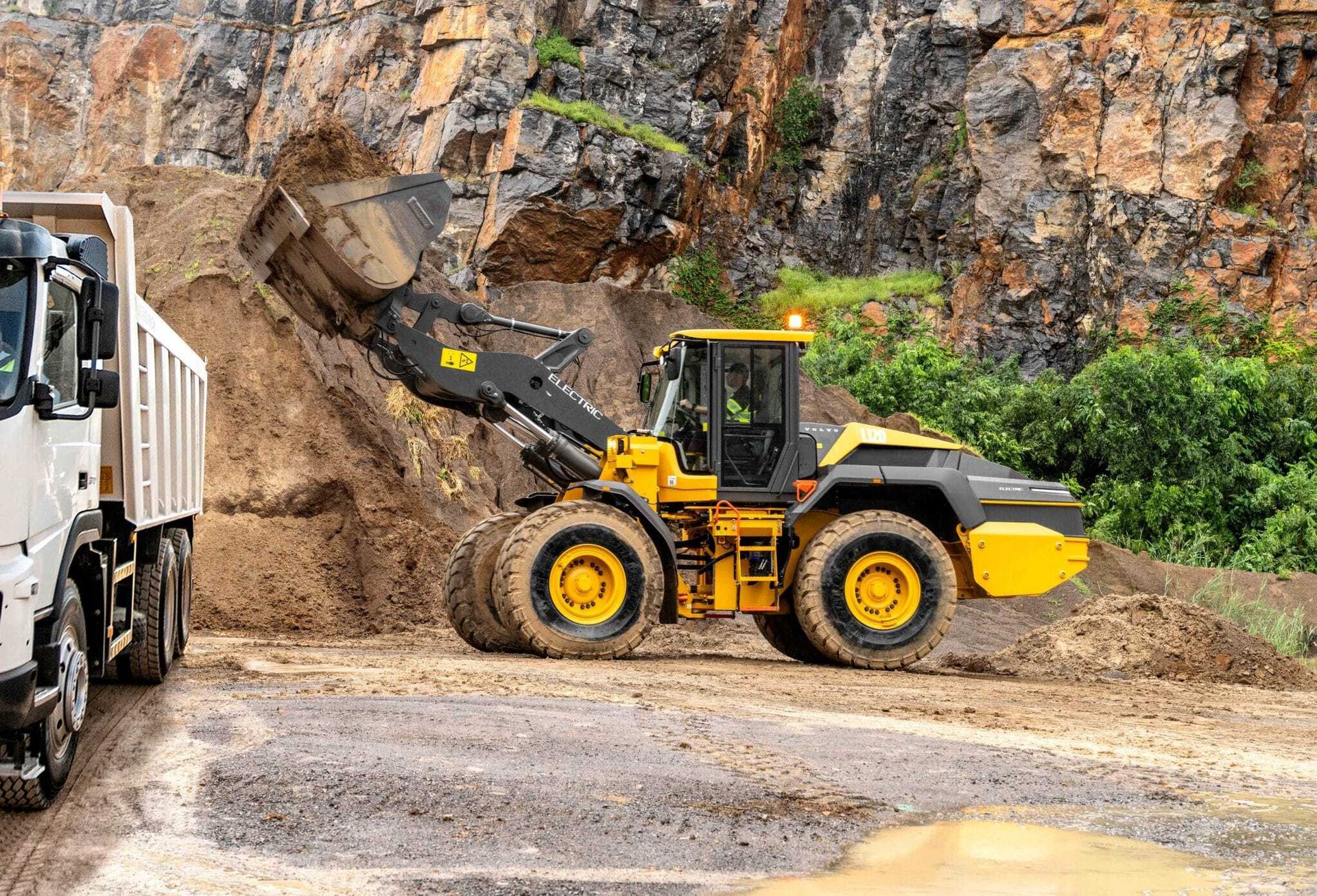 Yellow electric wheel loader filling a dump truck with dirt in a rocky quarry.
