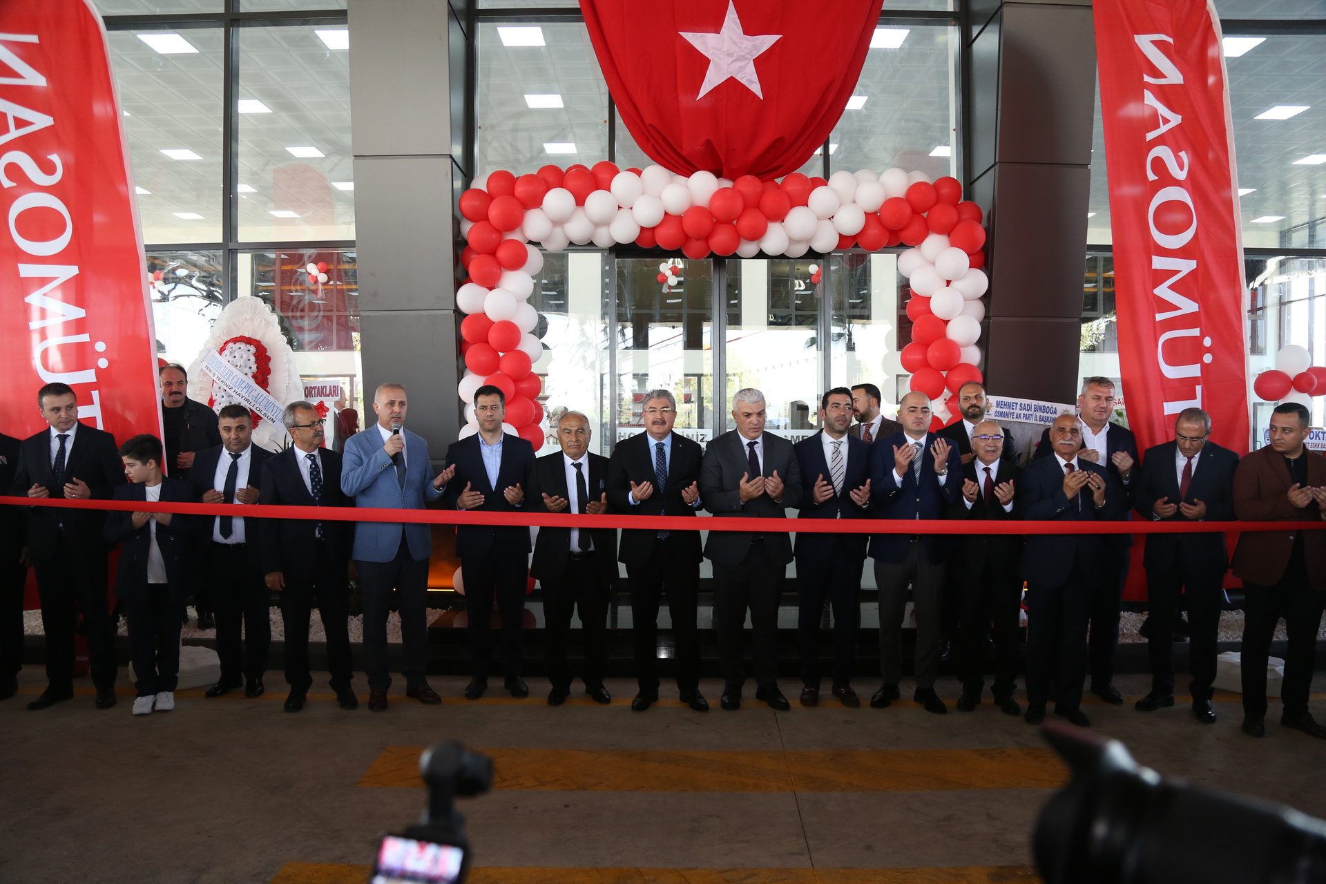 A group of men in suits participate in a ribbon-cutting ceremony, many with hands open in prayer, beneath a Turkish flag and red and white balloons.