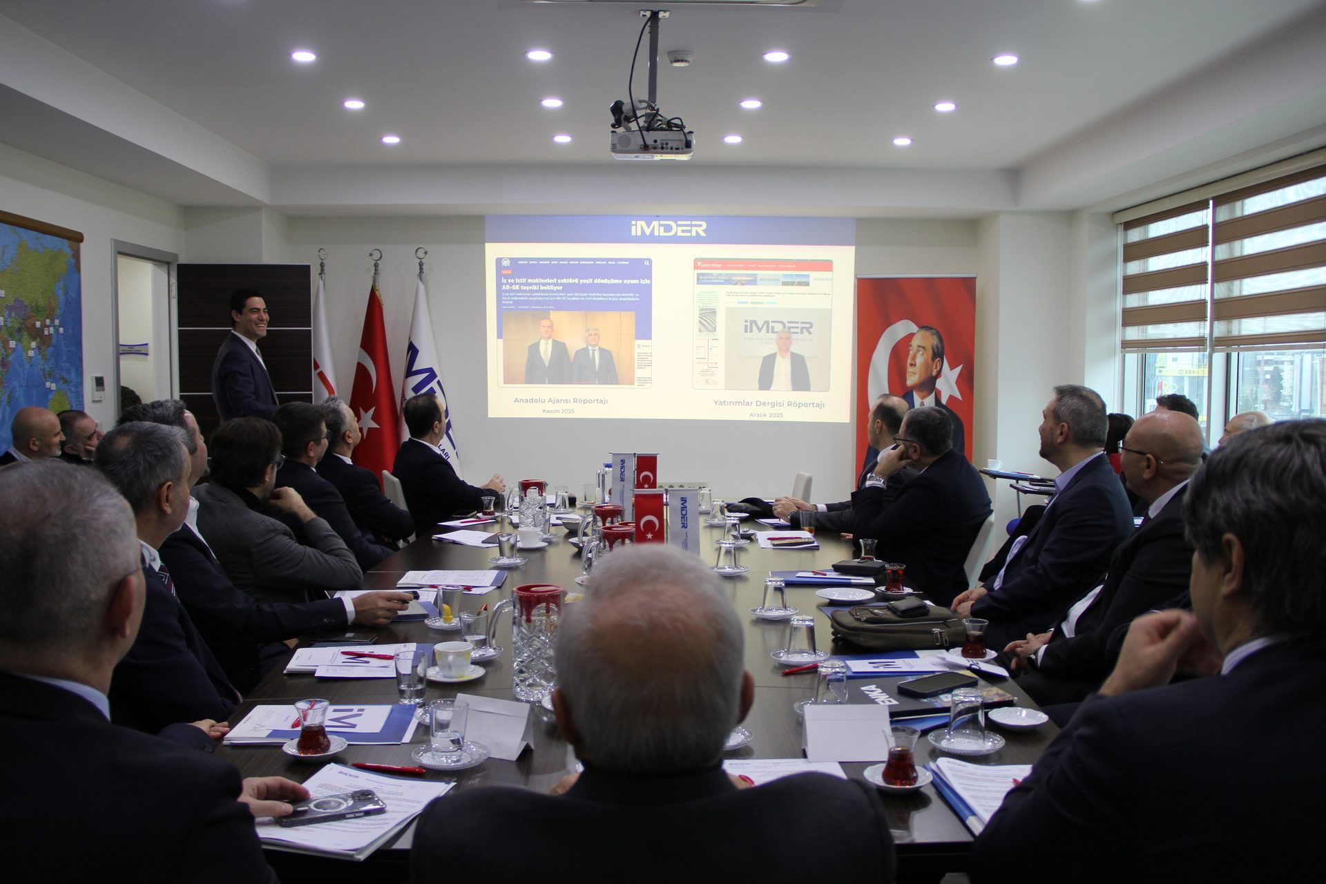 Men attend a business meeting around a table, viewing a presentation on a large projector screen.