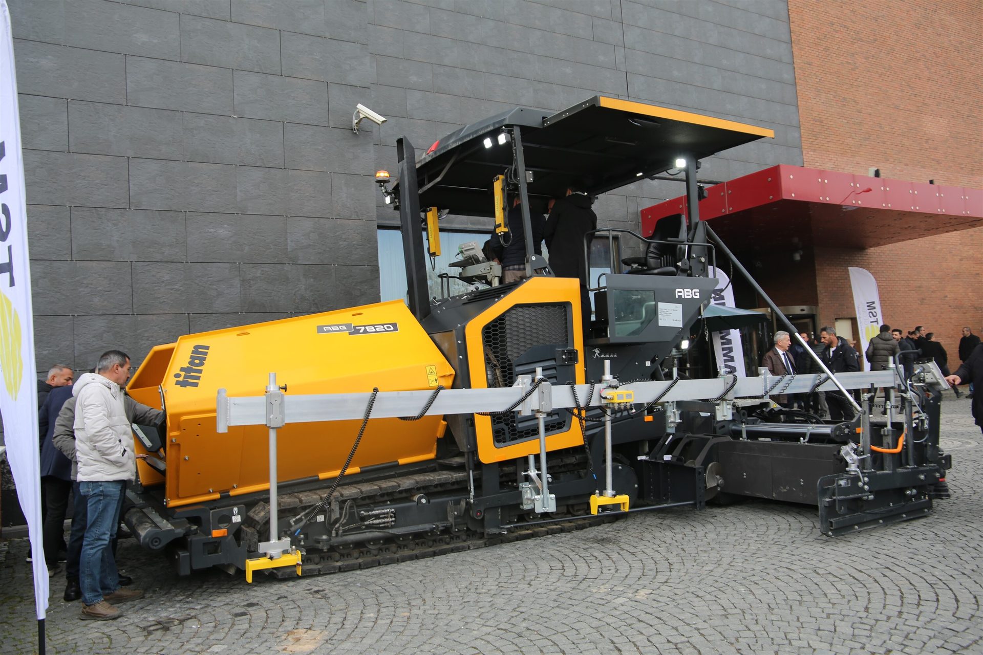 A large yellow and black ABG 7820 Titan asphalt paver, with people observing it at an outdoor display.