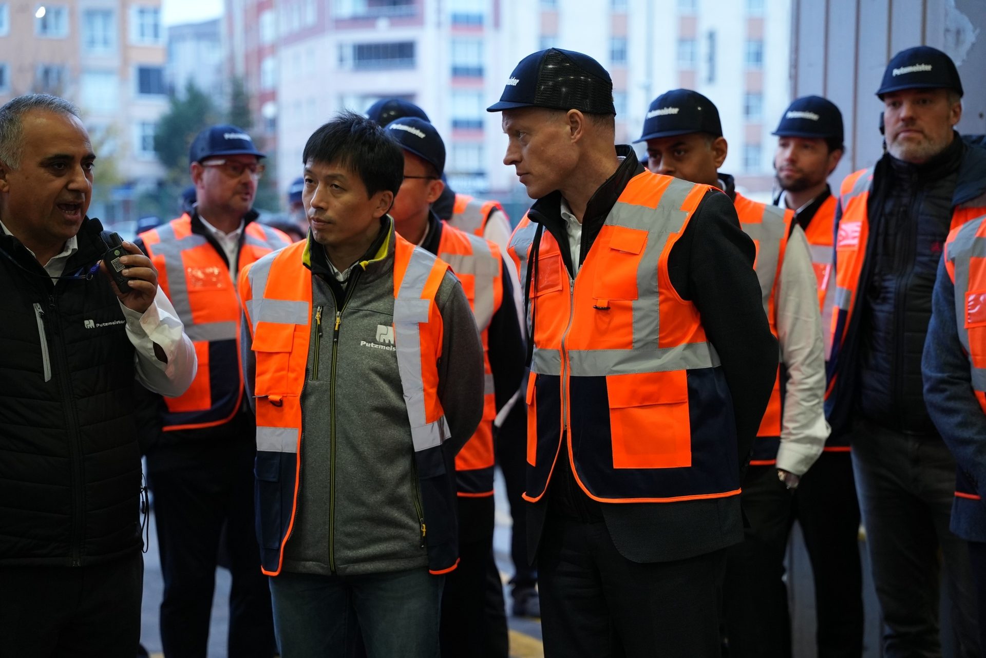 Men in orange safety vests and Putzmeister hats; one speaks into a device.