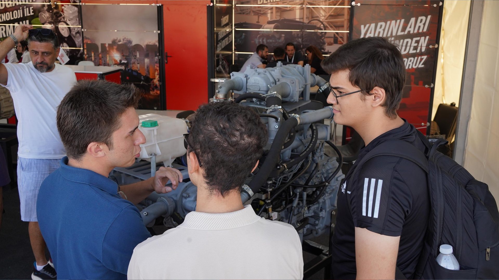 Three young men examining an engine display, with an adult man standing behind them.