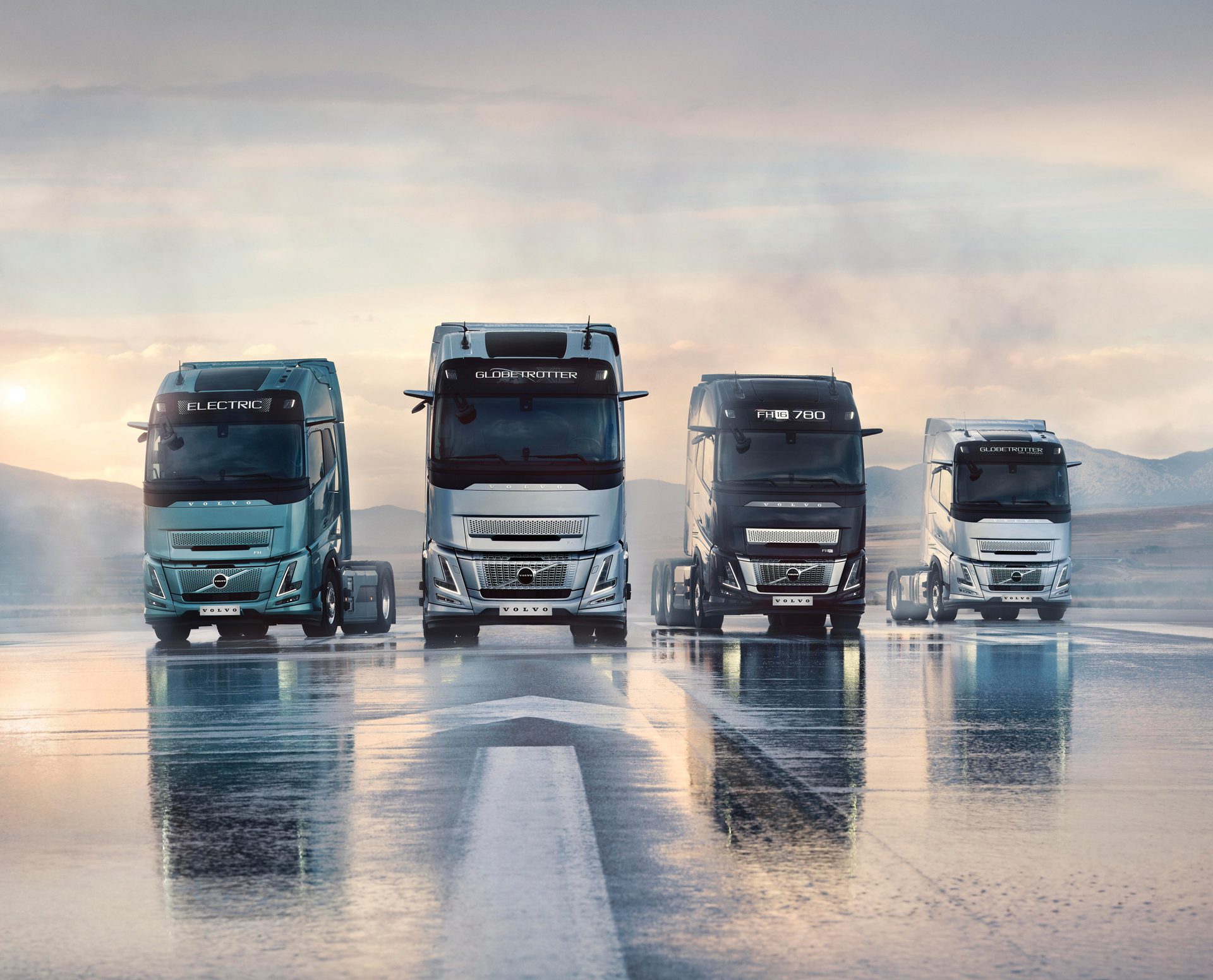 Four Volvo trucks, including an electric model, parked on a wet road with a sunrise/sunset in the background.