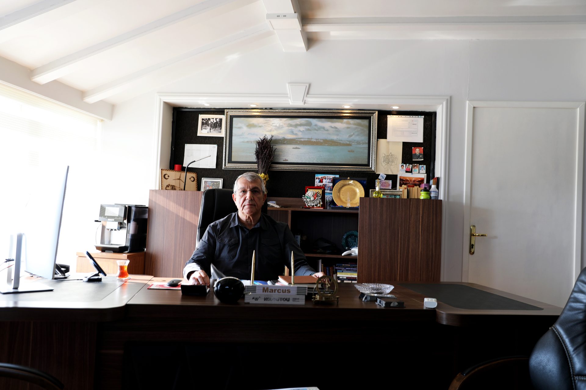 Older man with glasses at a dark office desk with a computer, coffee machine, and wall art.