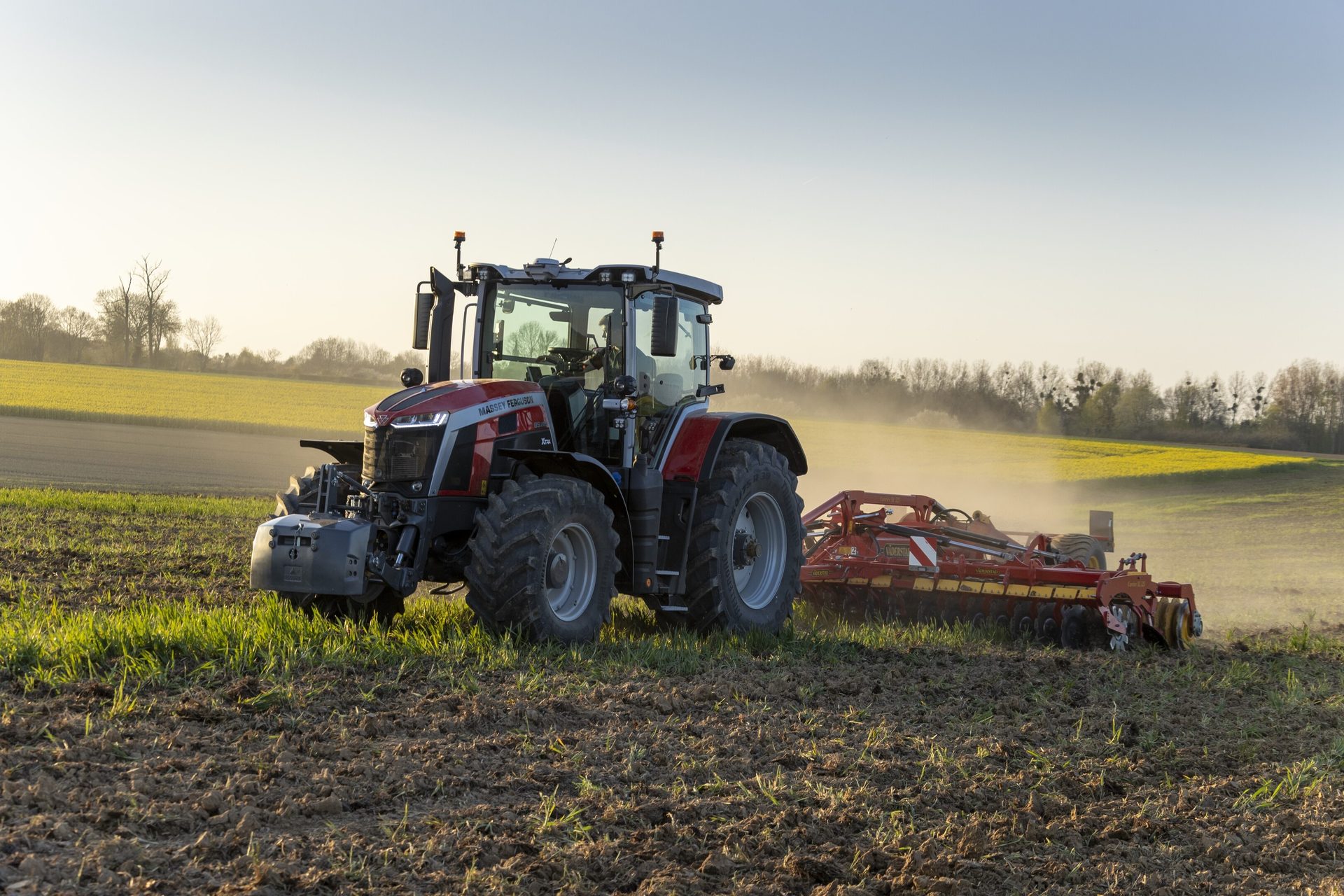 Red and grey tractor with an implement tilling a field at sunset, kicking up dust.