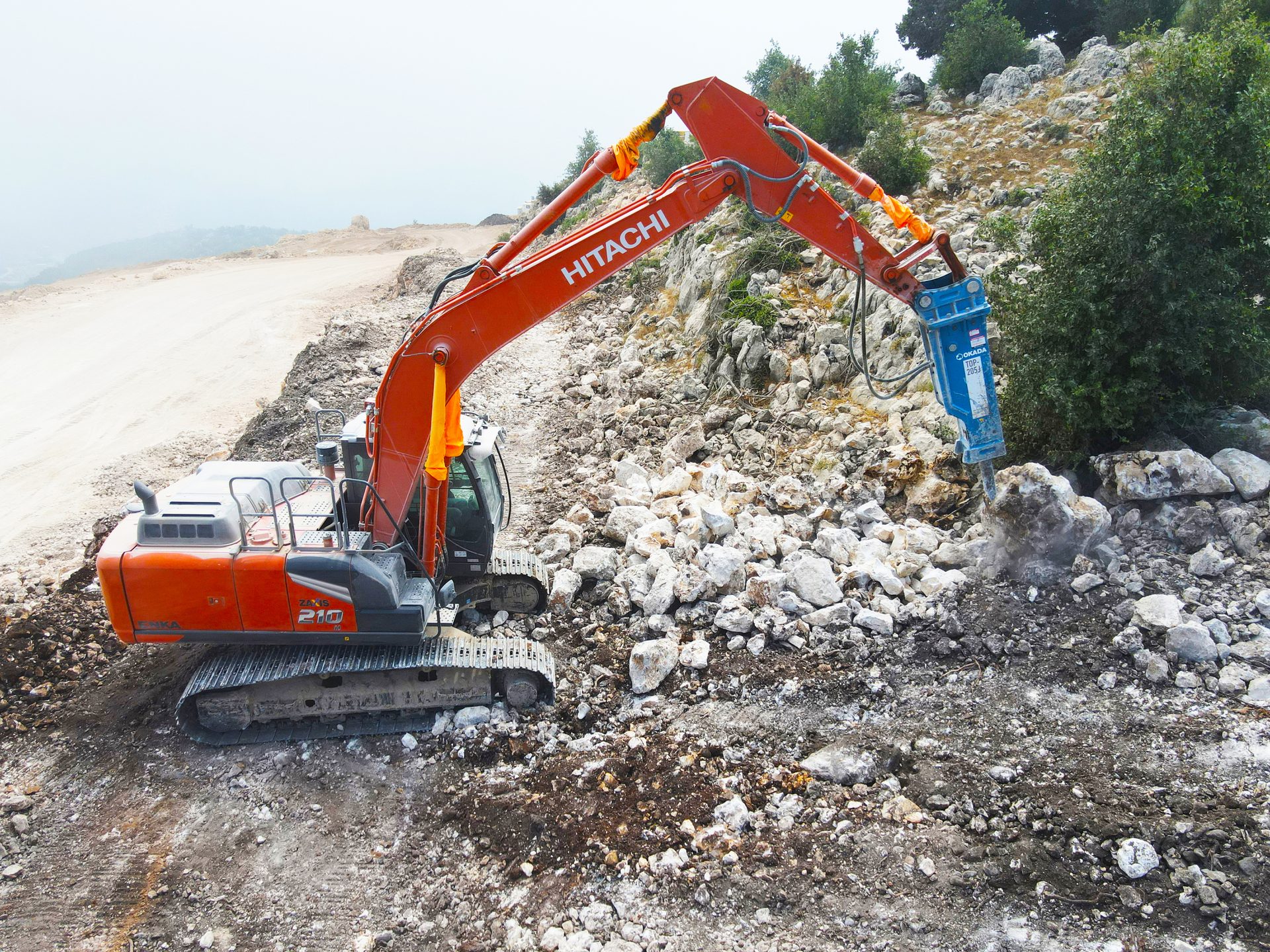An orange Hitachi excavator uses a hydraulic breaker to fragment rocks on a construction site hillside.