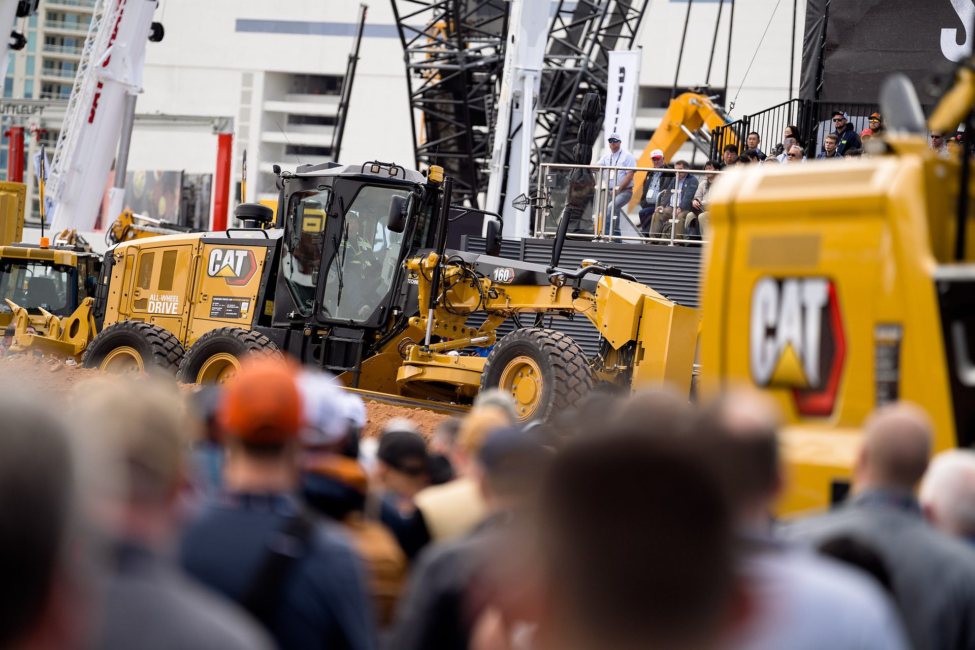 Yellow CAT motor grader 160 demo for crowd, with cranes in background.