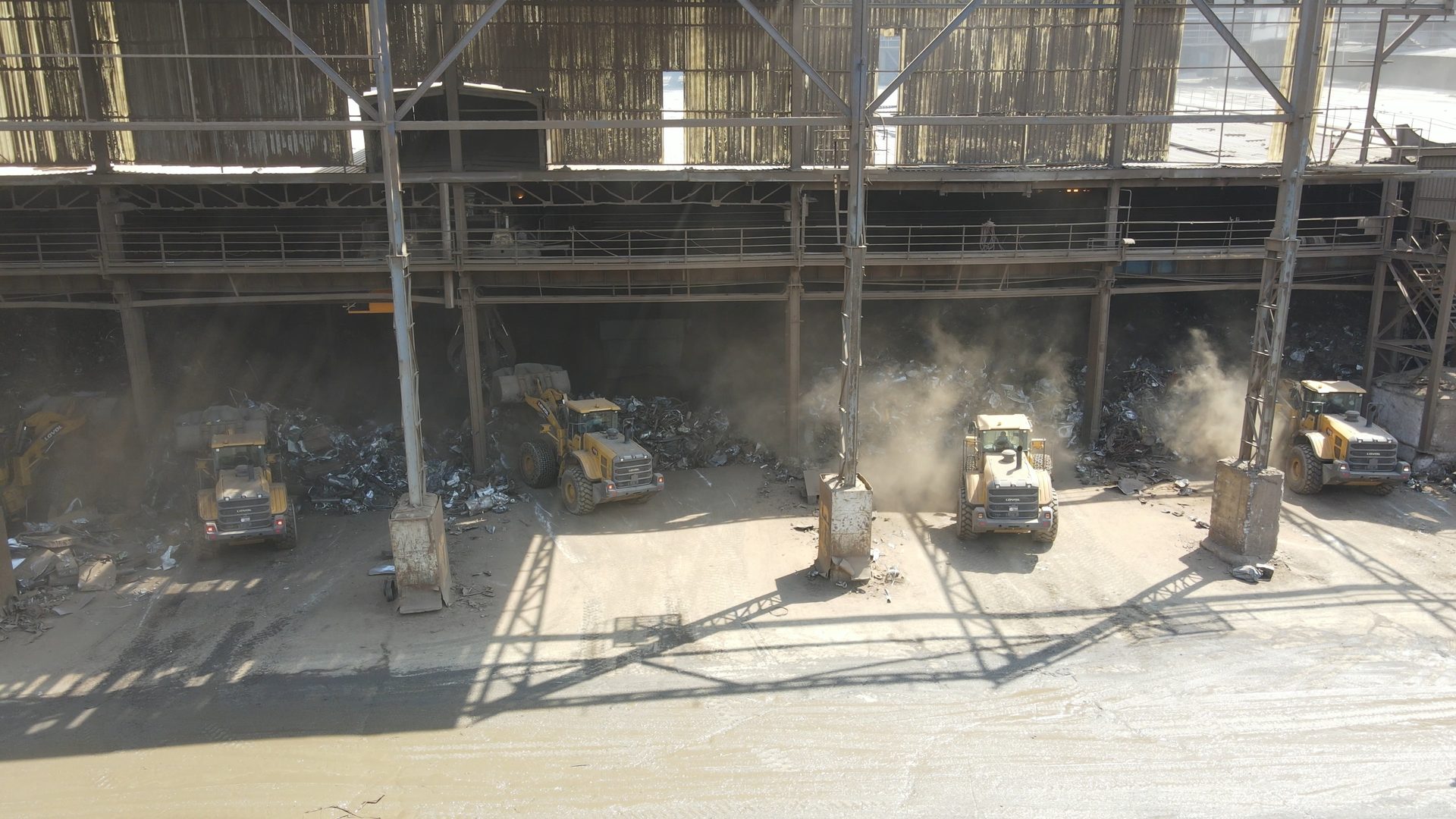 Dusty industrial scene with yellow heavy equipment moving large piles of scrap metal.