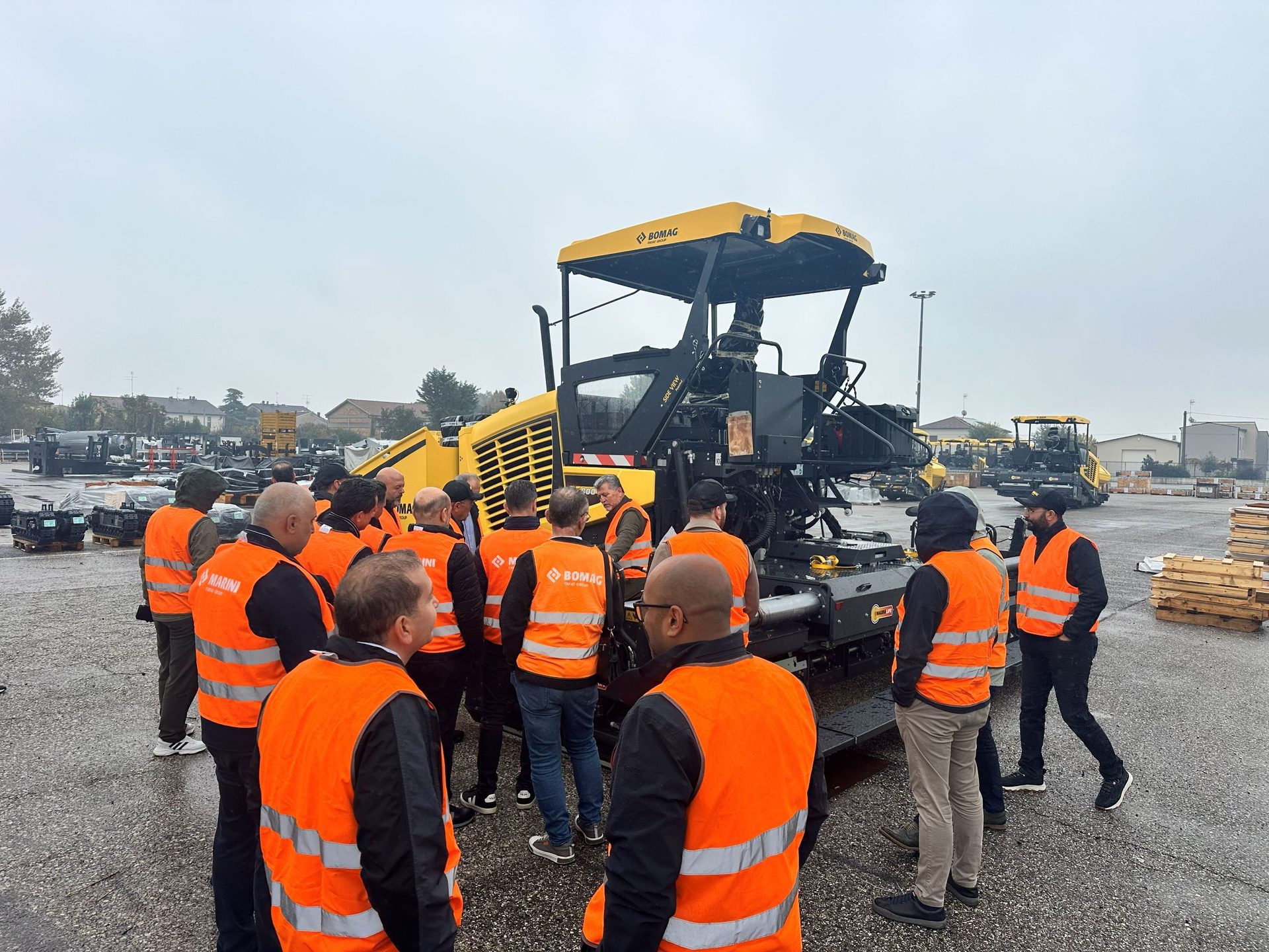 Group in orange safety vests gathered around a large yellow construction machine under an overcast sky.