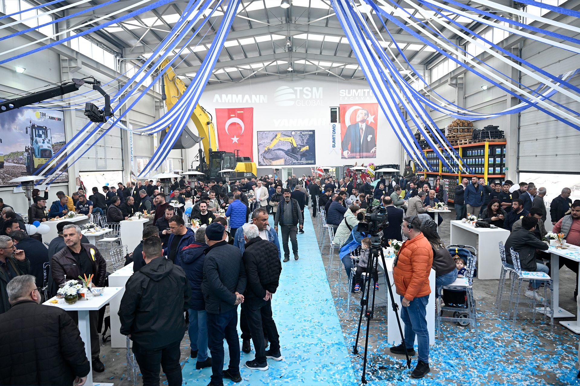 A large indoor gathering with many people, construction machinery, and blue and white decorations.