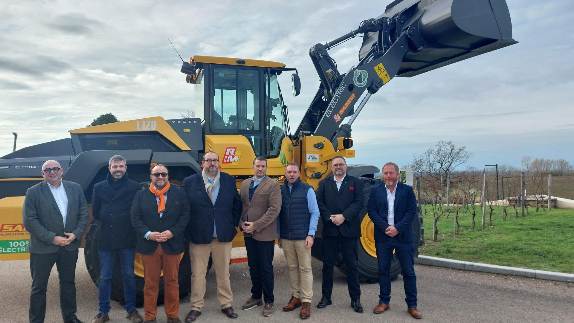 Eight men pose with a yellow Volvo L120 electric wheel loader in front of a vineyard.