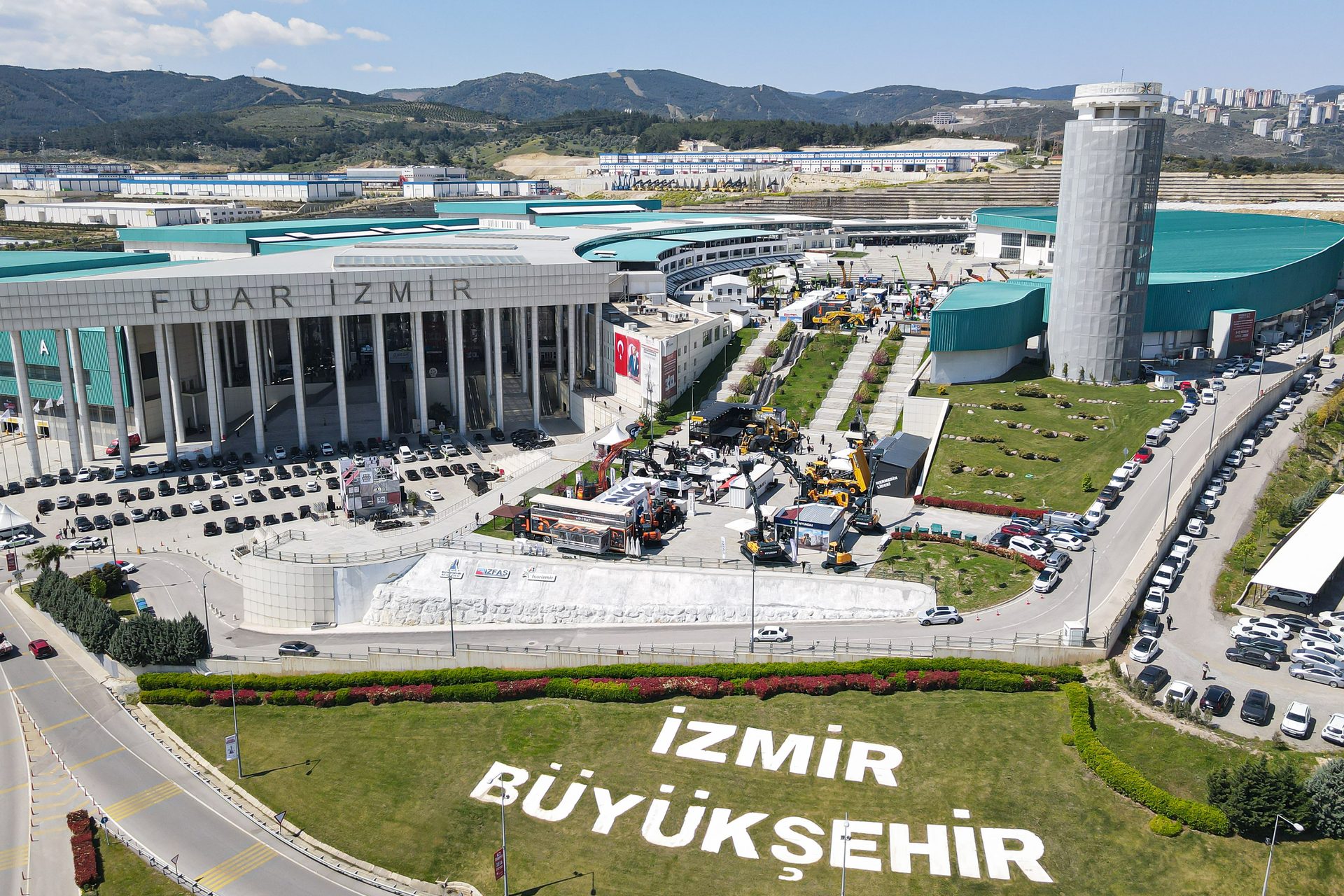 Aerial view of Fuar Izmir exhibition center with outdoor construction equipment displays and 'IZMIR BÜYÜKŞEHİR' on the lawn.
