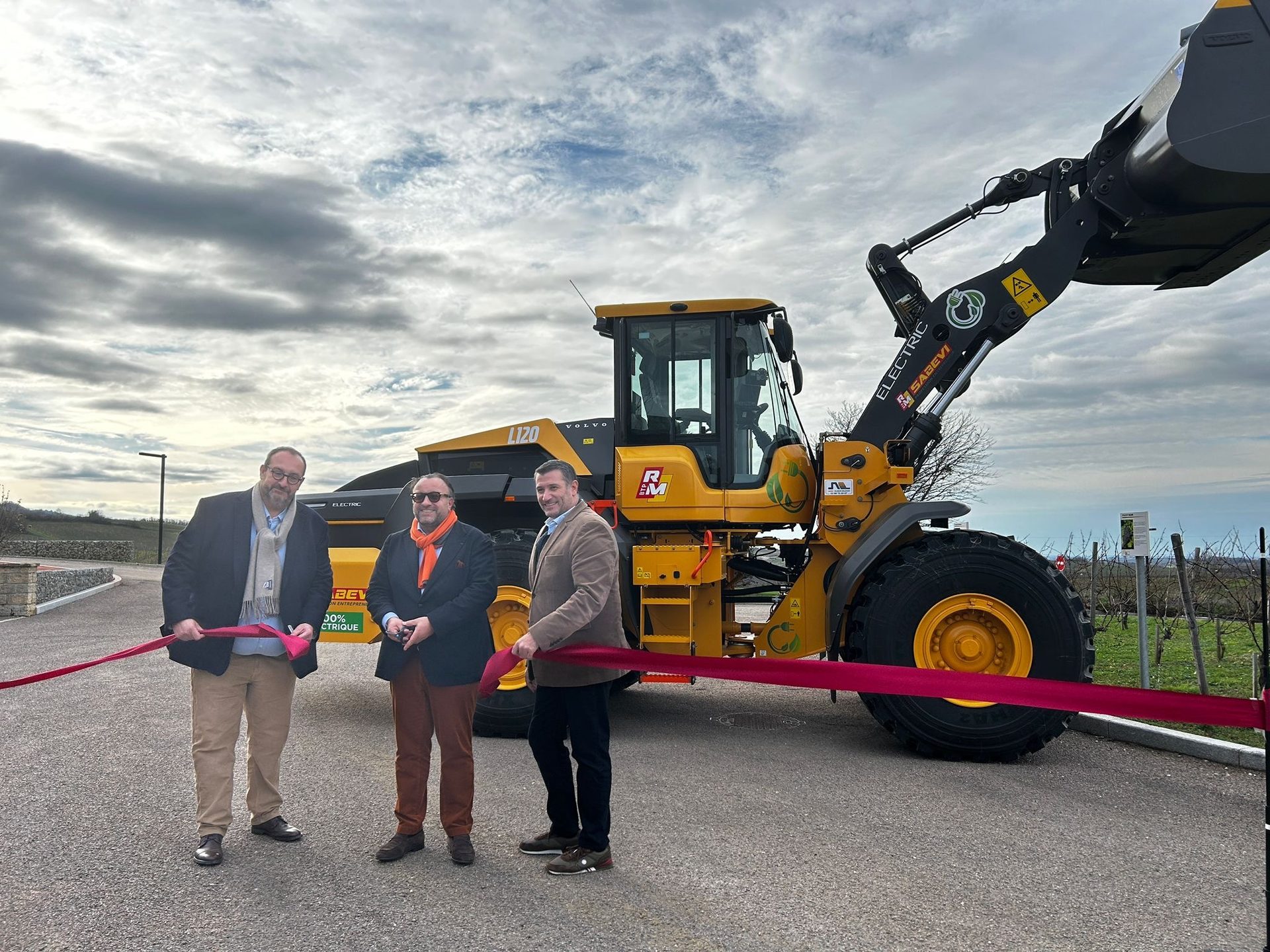 Three men cut a red ribbon in front of a yellow electric Volvo L120 loader at an outdoor ceremony.