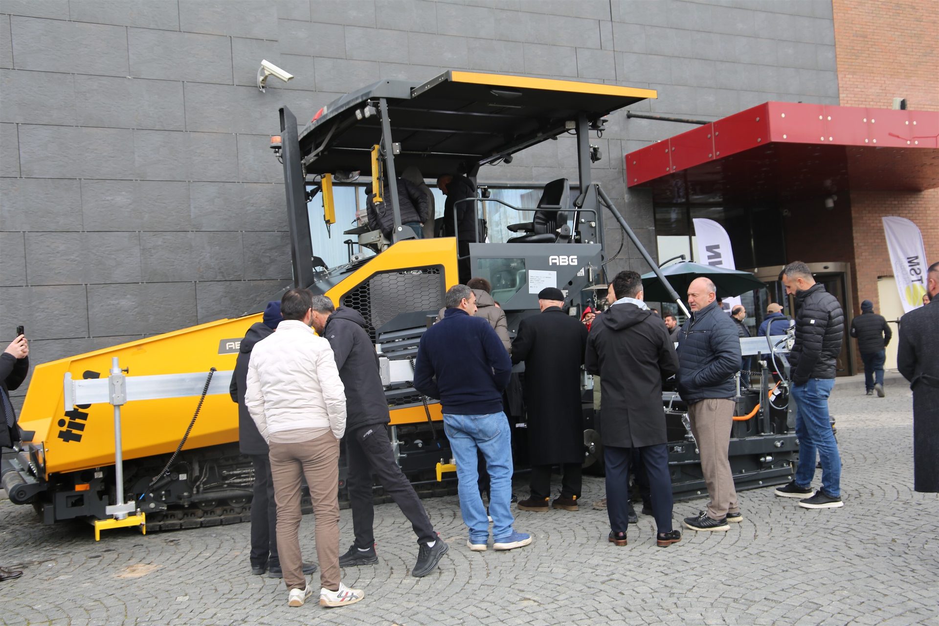 Men gathered around a large yellow and black paving machine, inspecting it at an outdoor event.