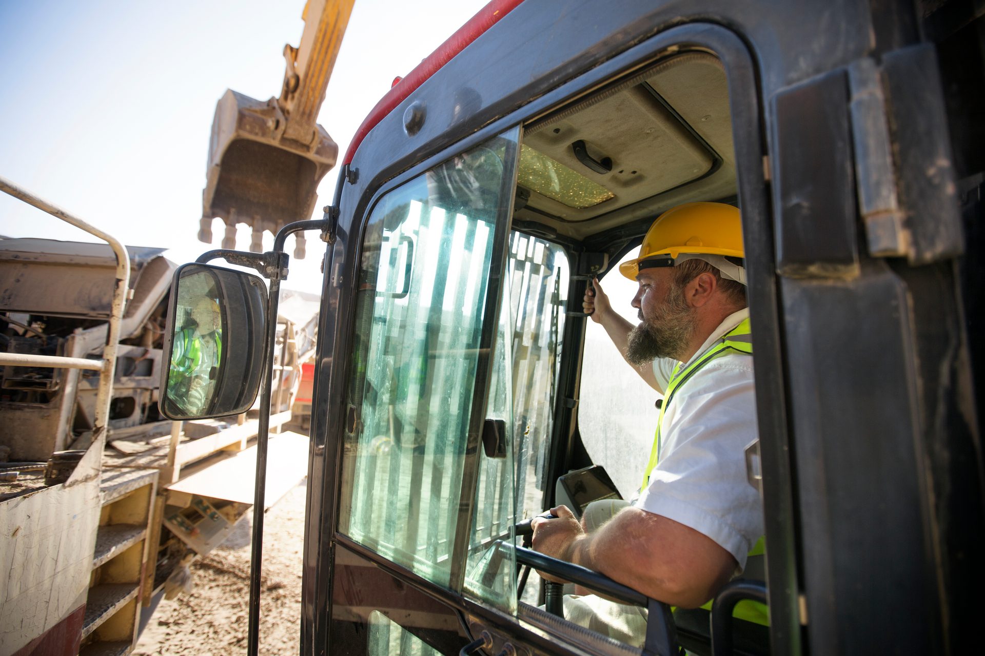 Bearded man in yellow hard hat and safety vest operating excavator from cab.