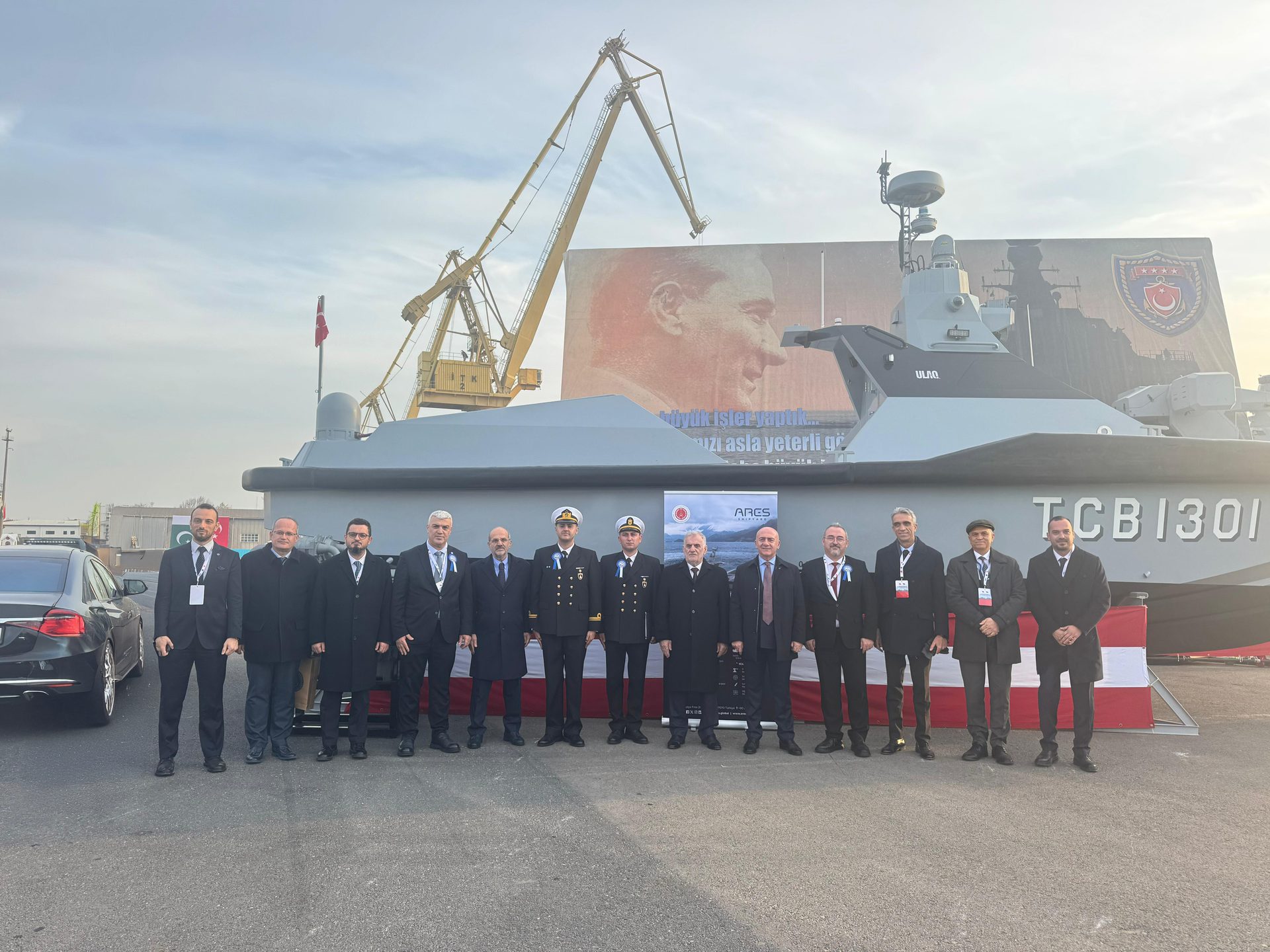 Men in suits and naval uniforms pose with the ULAQ patrol boat, featuring Atatürk's image and cranes.