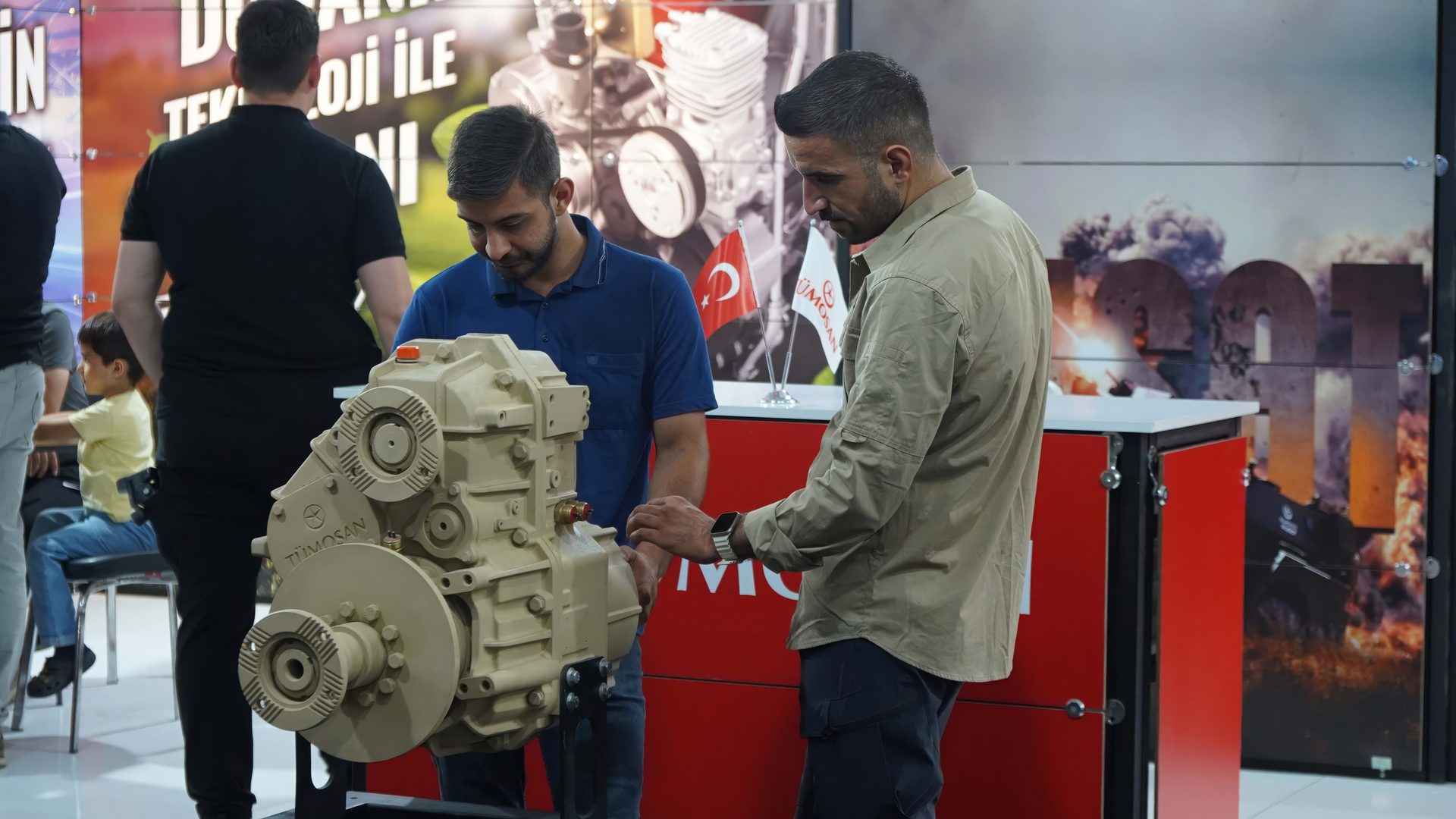 Two men examine a TÜMOSAN engine at an exhibition booth with Turkish flags.