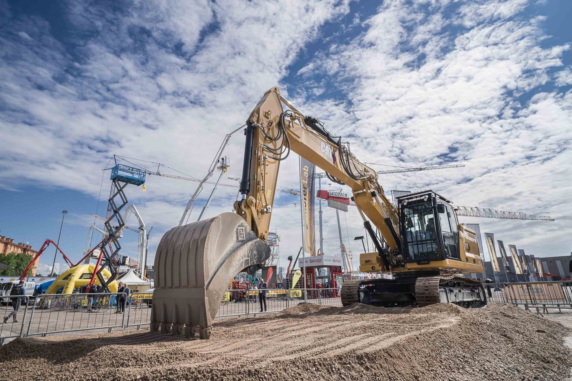 A large yellow CAT excavator on a dirt mound at an outdoor construction exhibition under a cloudy sky.