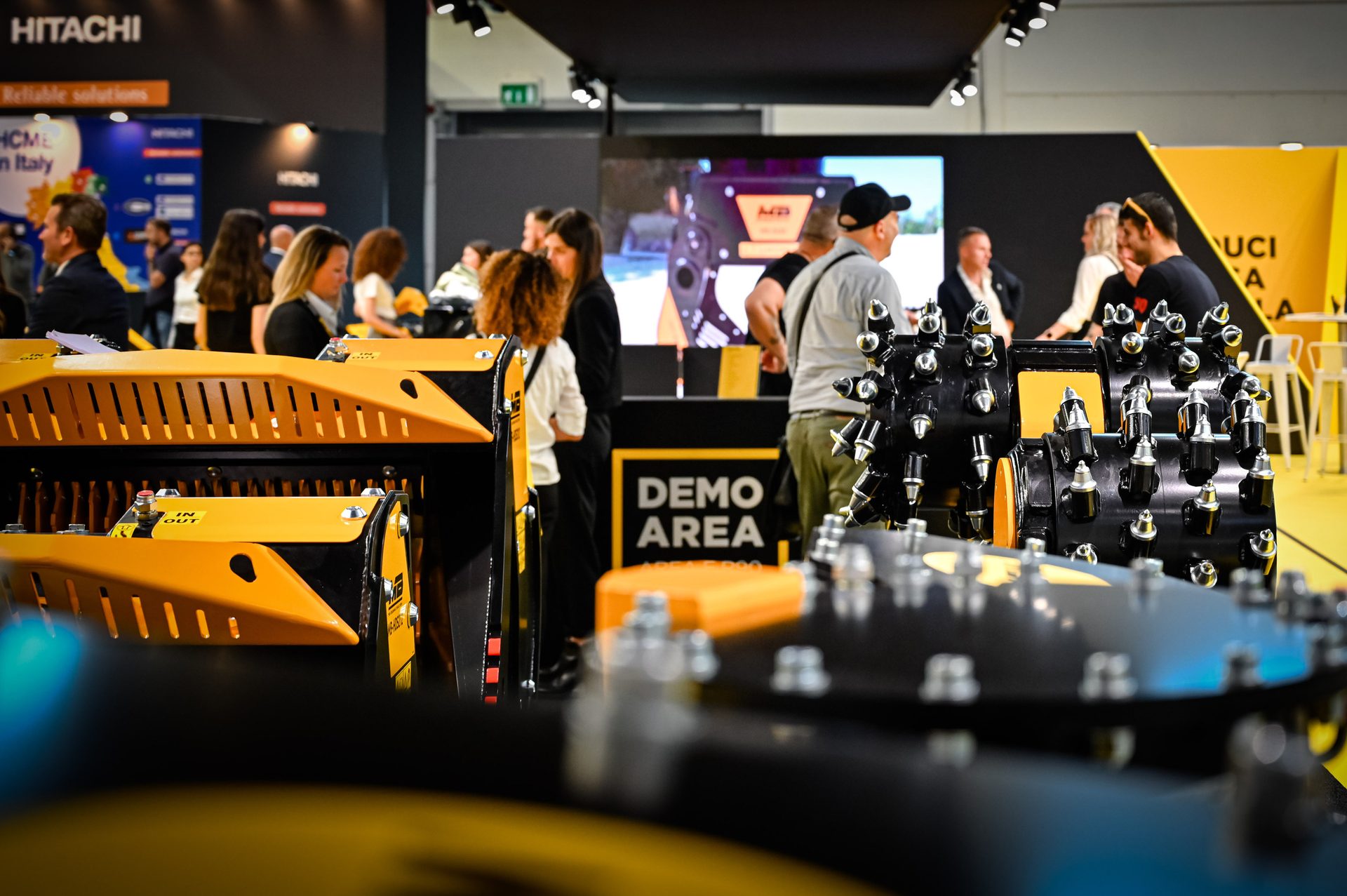 Trade show: people view yellow/black industrial machinery. Hitachi and 'Demo Area' signs.